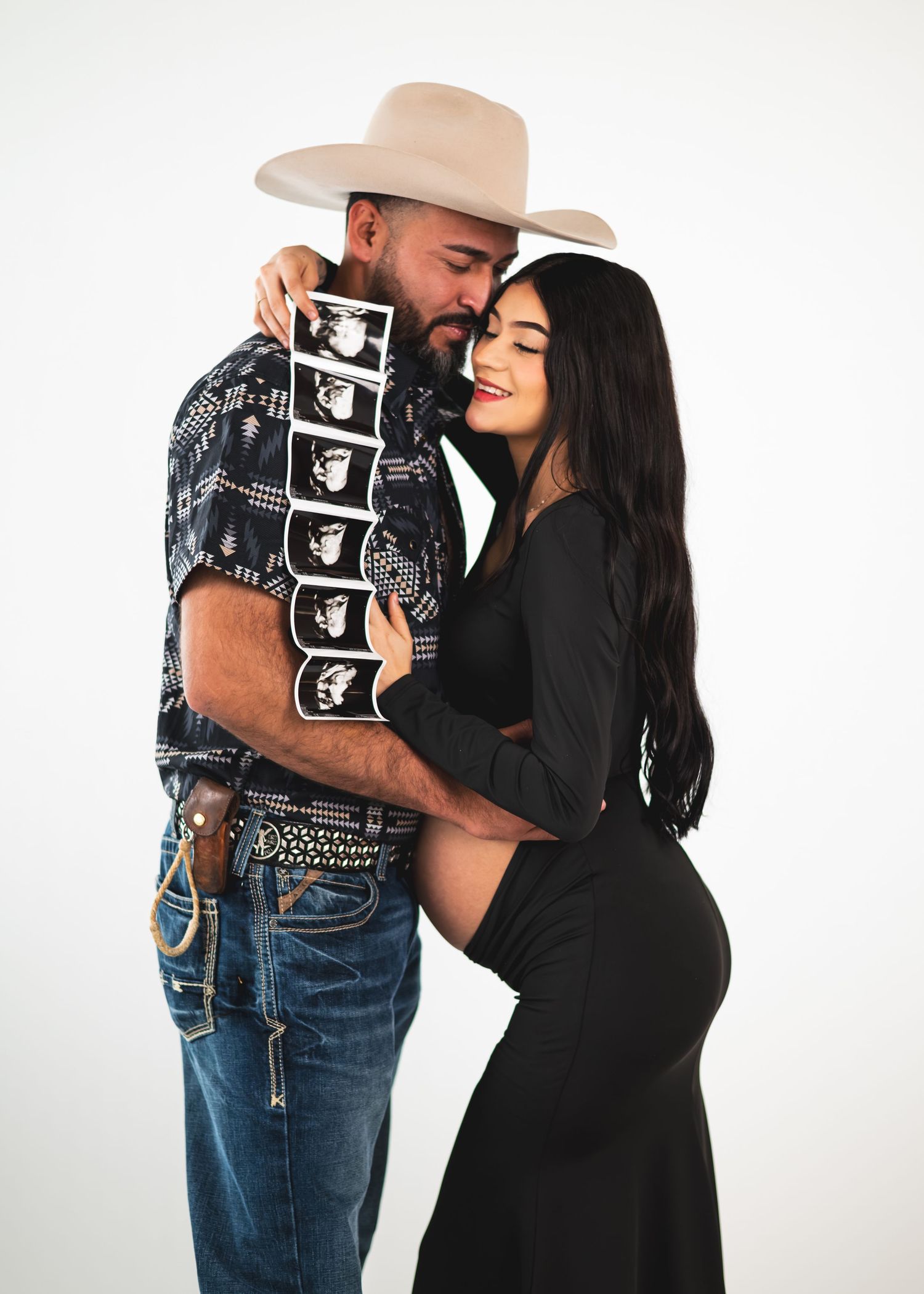 Couple in maternity photo shoot holding ultrasound strip, man in cowboy hat and woman in black dress showing baby bump.