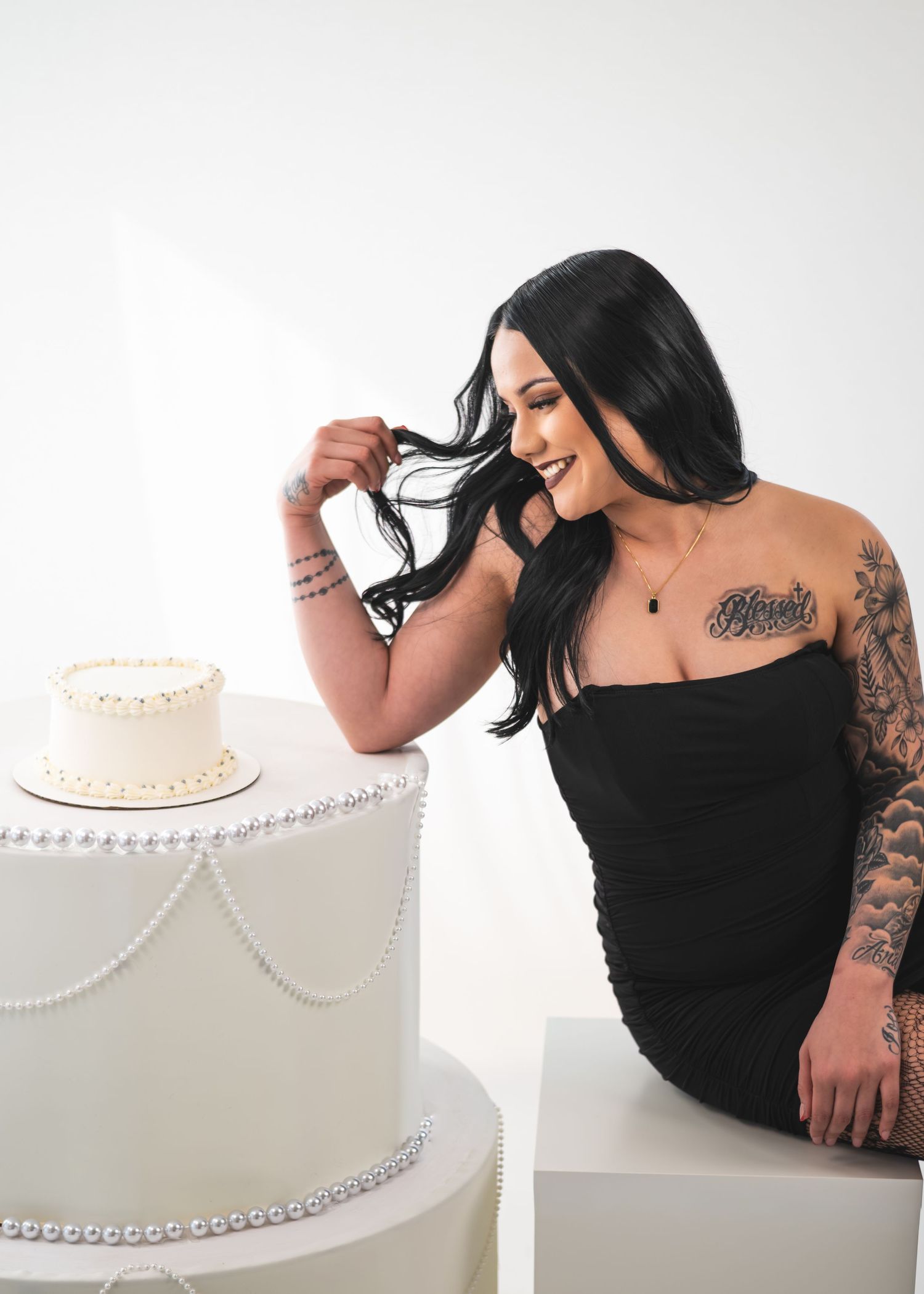 Woman in black dress with tattoos smiles beside elegant white tiered wedding cake with pearl details.