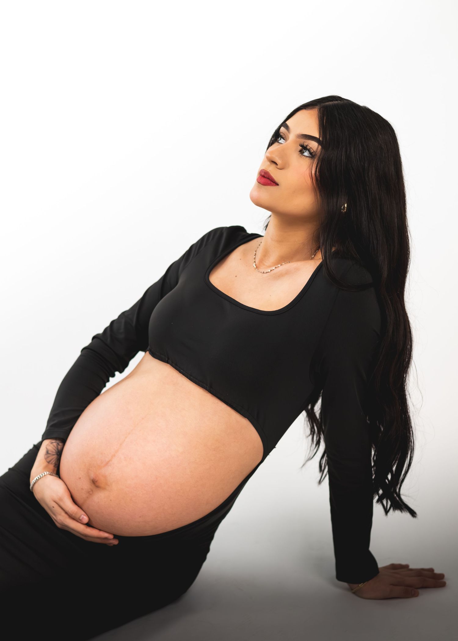 Pregnant woman in black crop top and skirt cradling baby bump, long dark hair, red lips, studio white background.