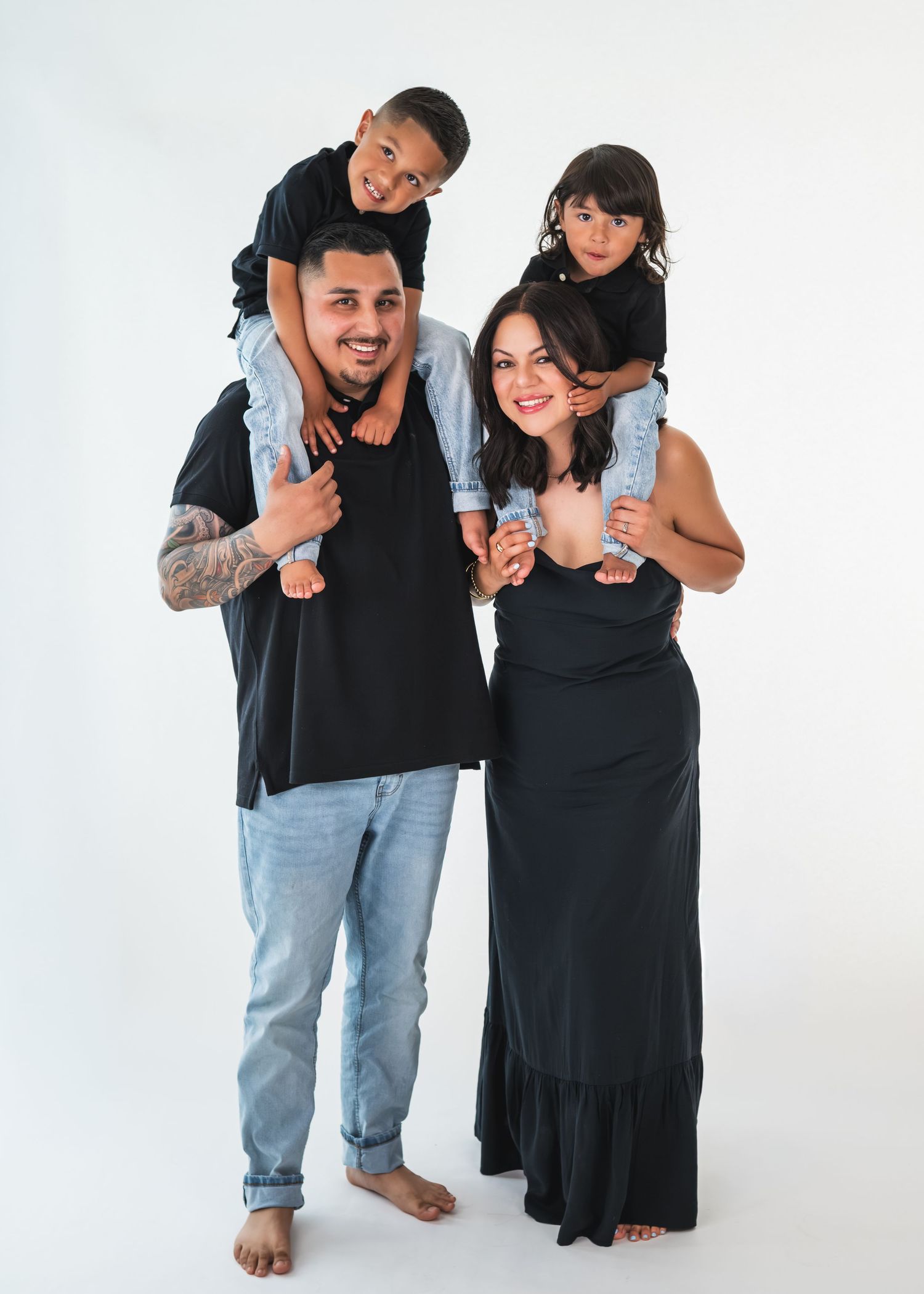 Family portrait of parents carrying two young children on their shoulders, all dressed in black, against a white background.