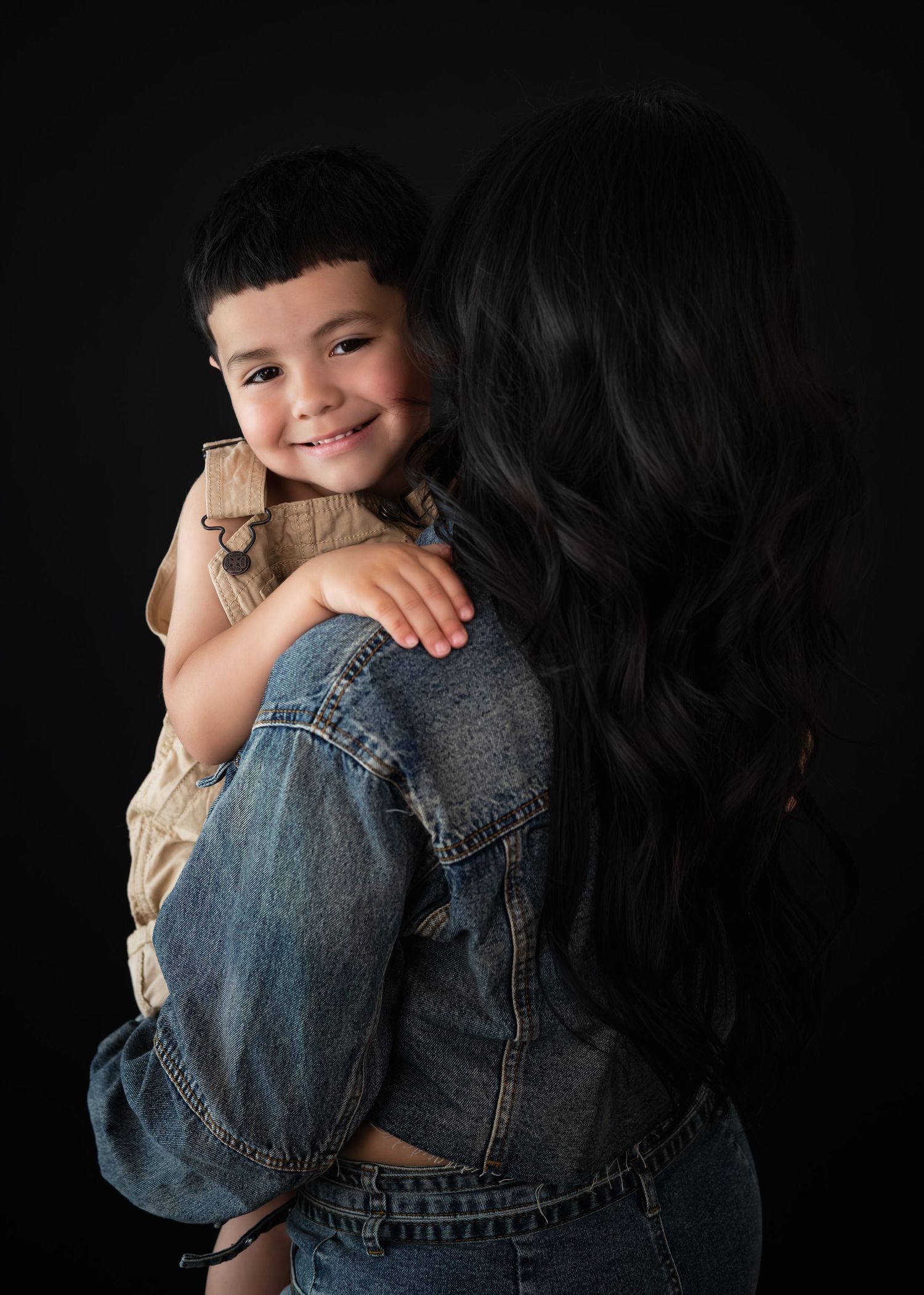 A smiling young boy being held by a woman with long dark hair in a denim jacket against a black background.