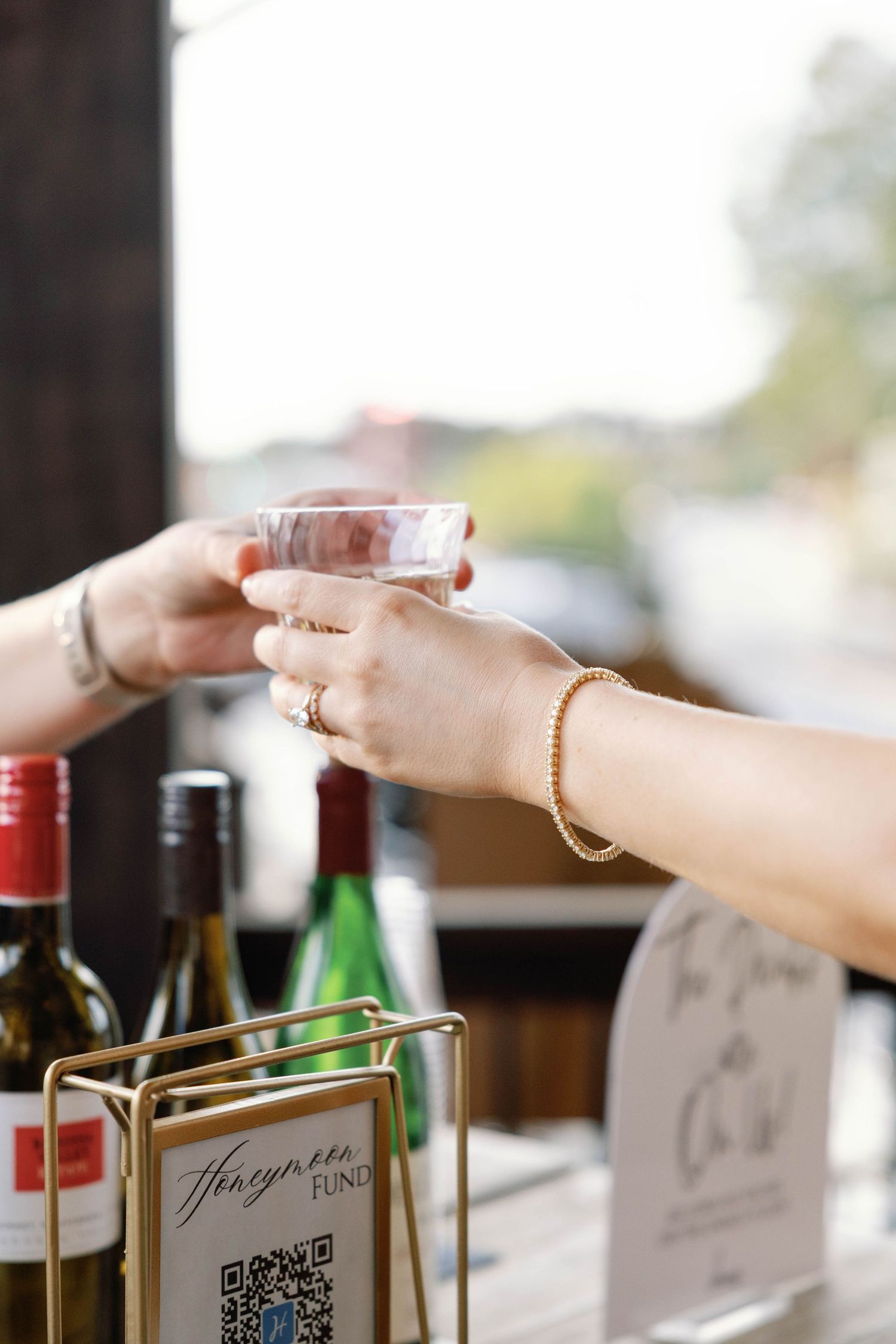 Hands holding a glass of wine at a tasting event with wine bottles and QR code menus visible in the background.