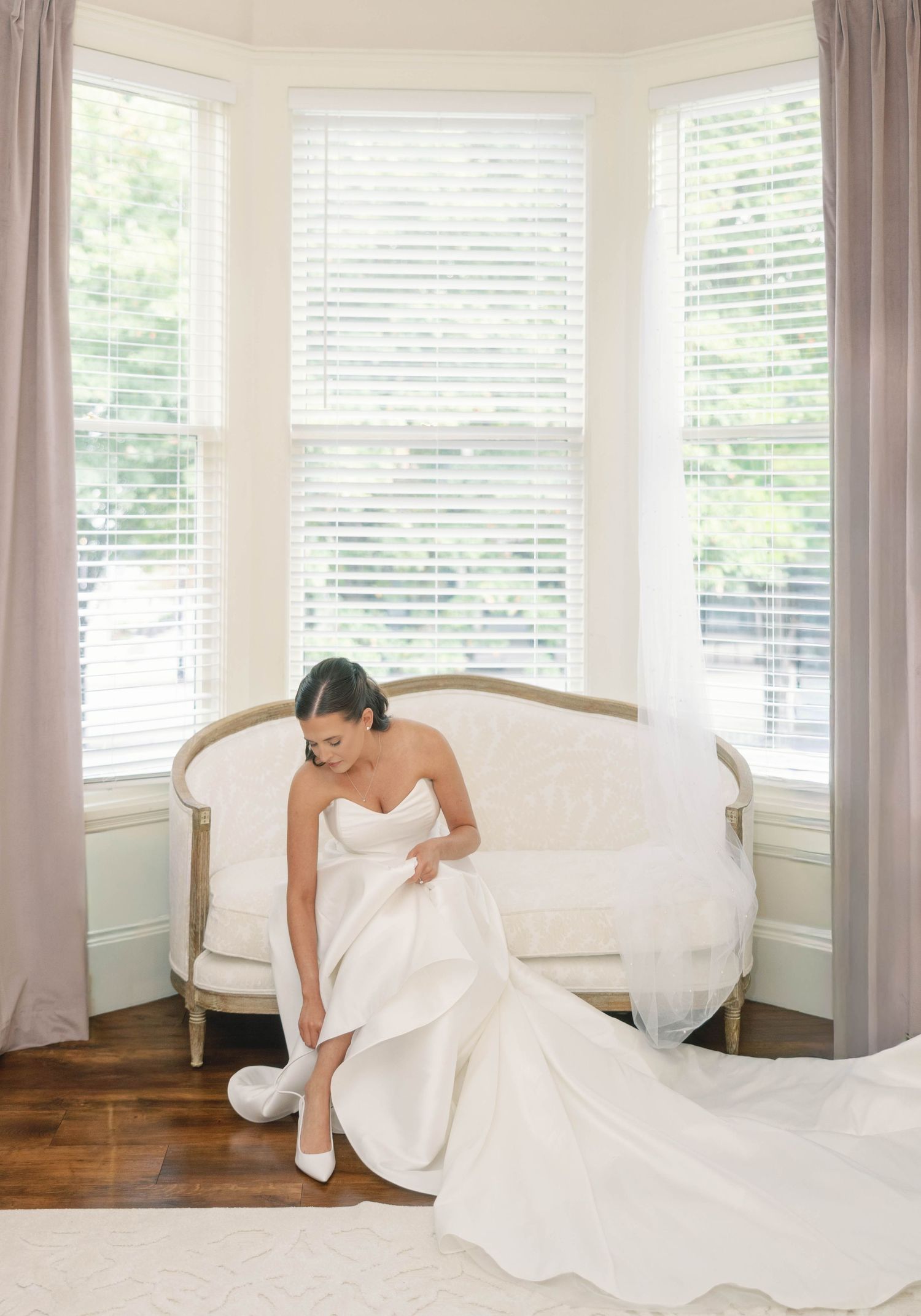 A strapless white wedding dress being adjusted in an elegant bay window setting with hardwood floors.