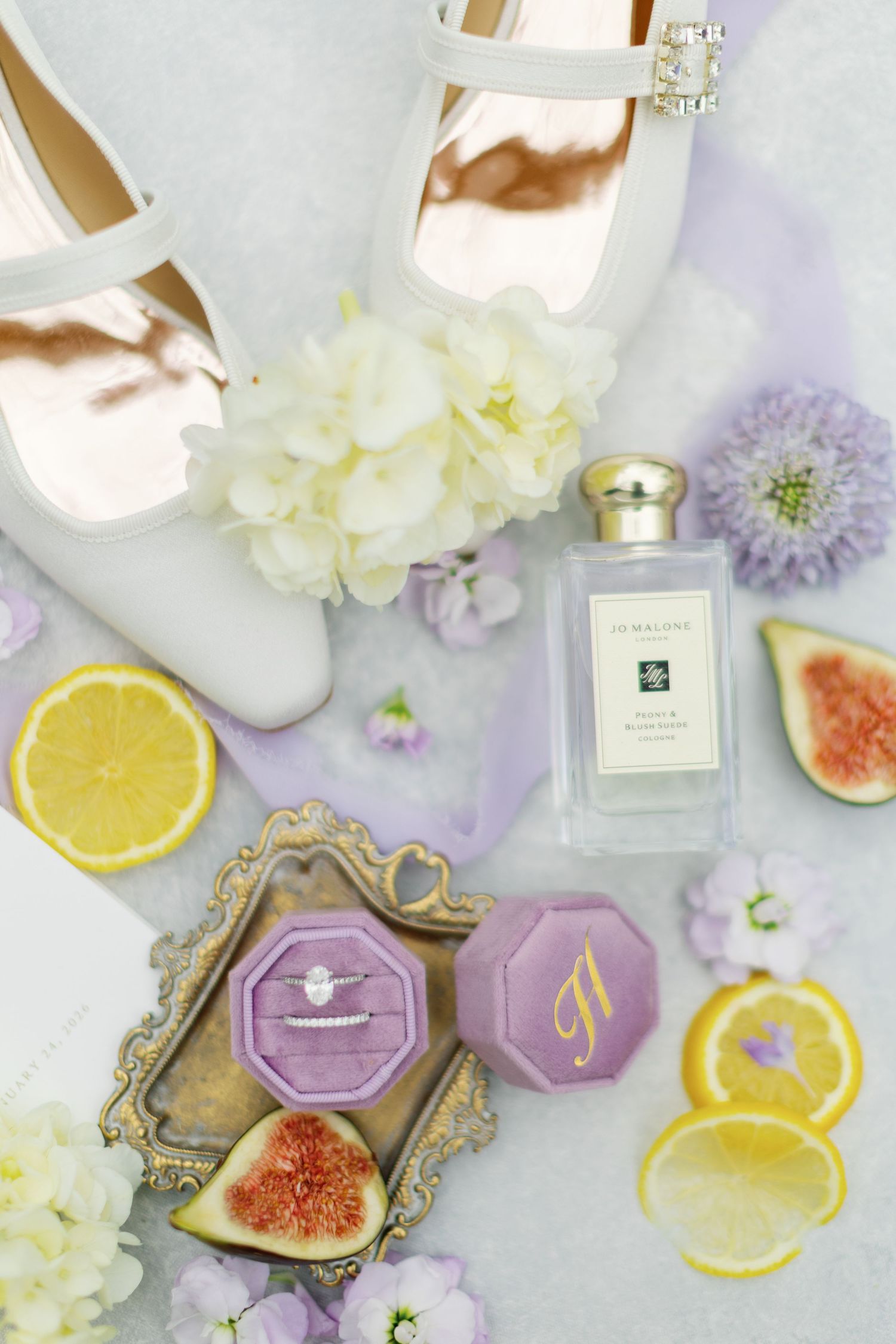 Bridal flat lay with white heels, Jo Malone perfume, purple ring box, engagement ring, figs, lemon slices, and flowers.