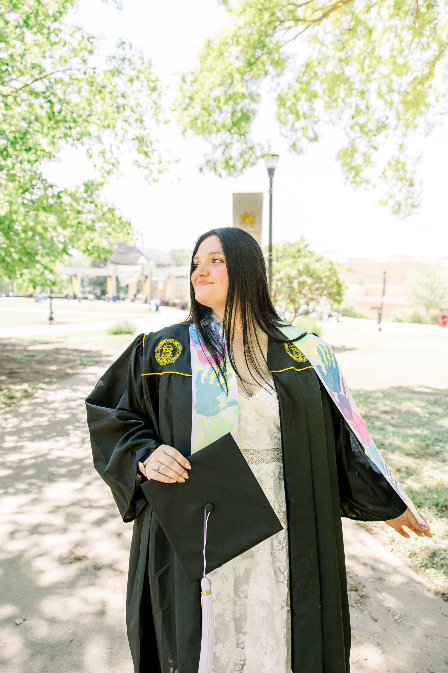 Graduate in black academic regalia holds diploma on sunny campus pathway.