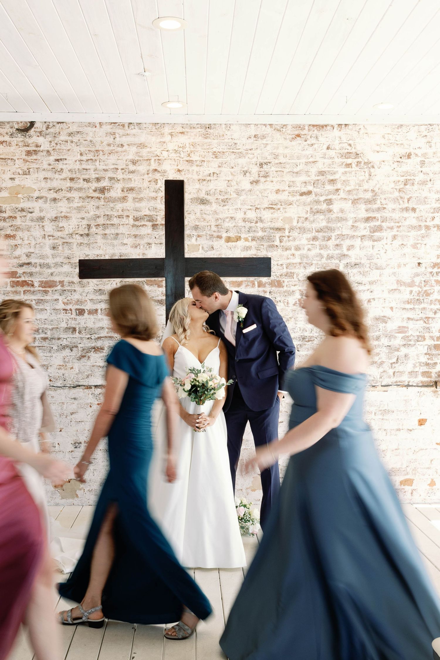 Wedding party dances in front of a rustic brick wall and black cross in a modern industrial venue.