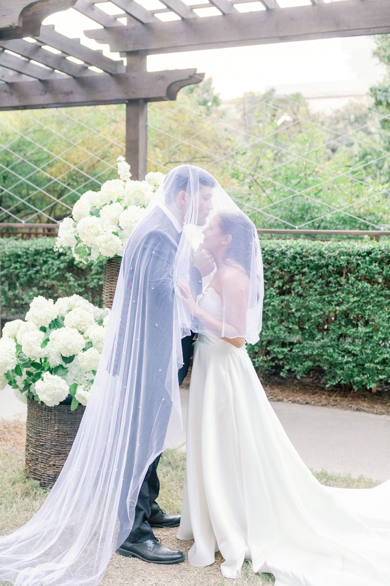 A romantic wedding moment under a pergola with flowing white veil and hydrangea flowers in an outdoor garden setting.