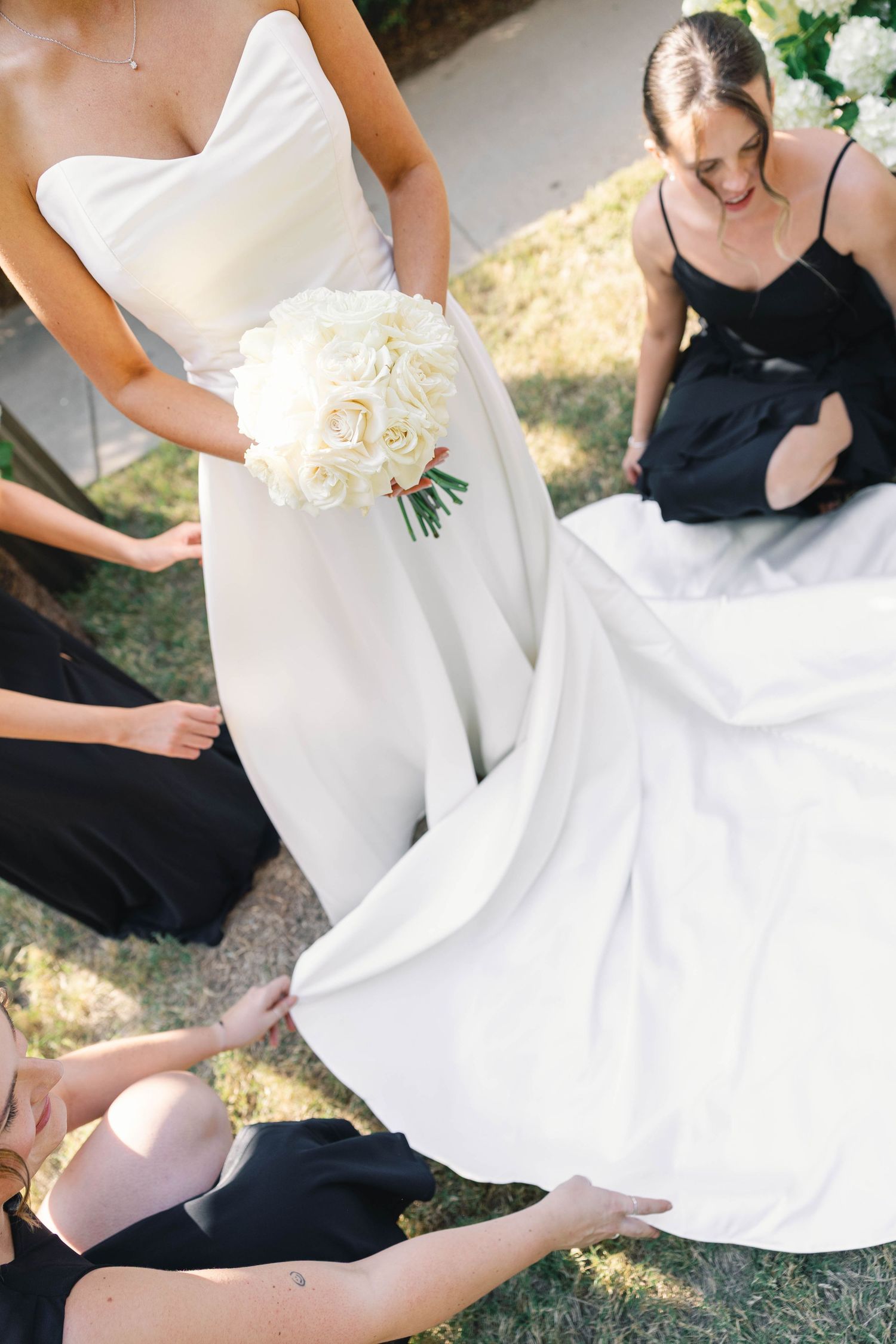 Wedding party members in black dresses helping adjust the train of an elegant white bridal gown outdoors.