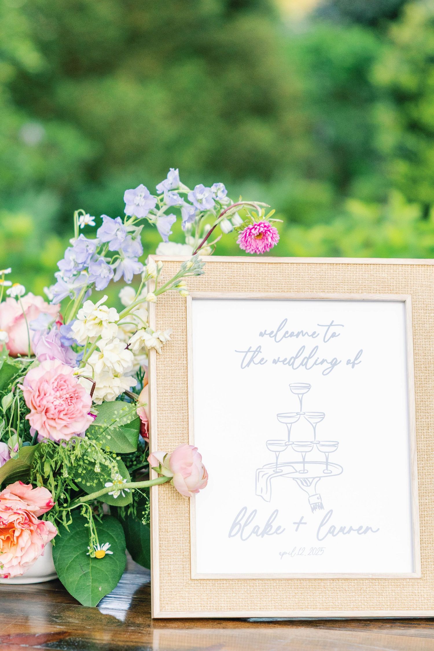 Wedding welcome sign in gold frame surrounded by pink and purple floral arrangements.