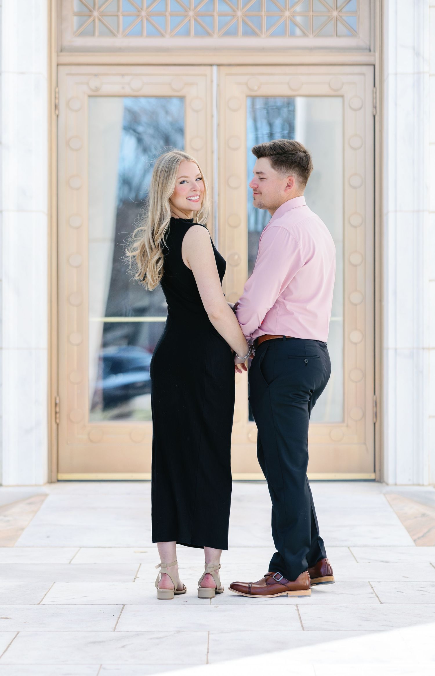 Stylish couple in black dress and pink shirt pose together at ornate entrance.