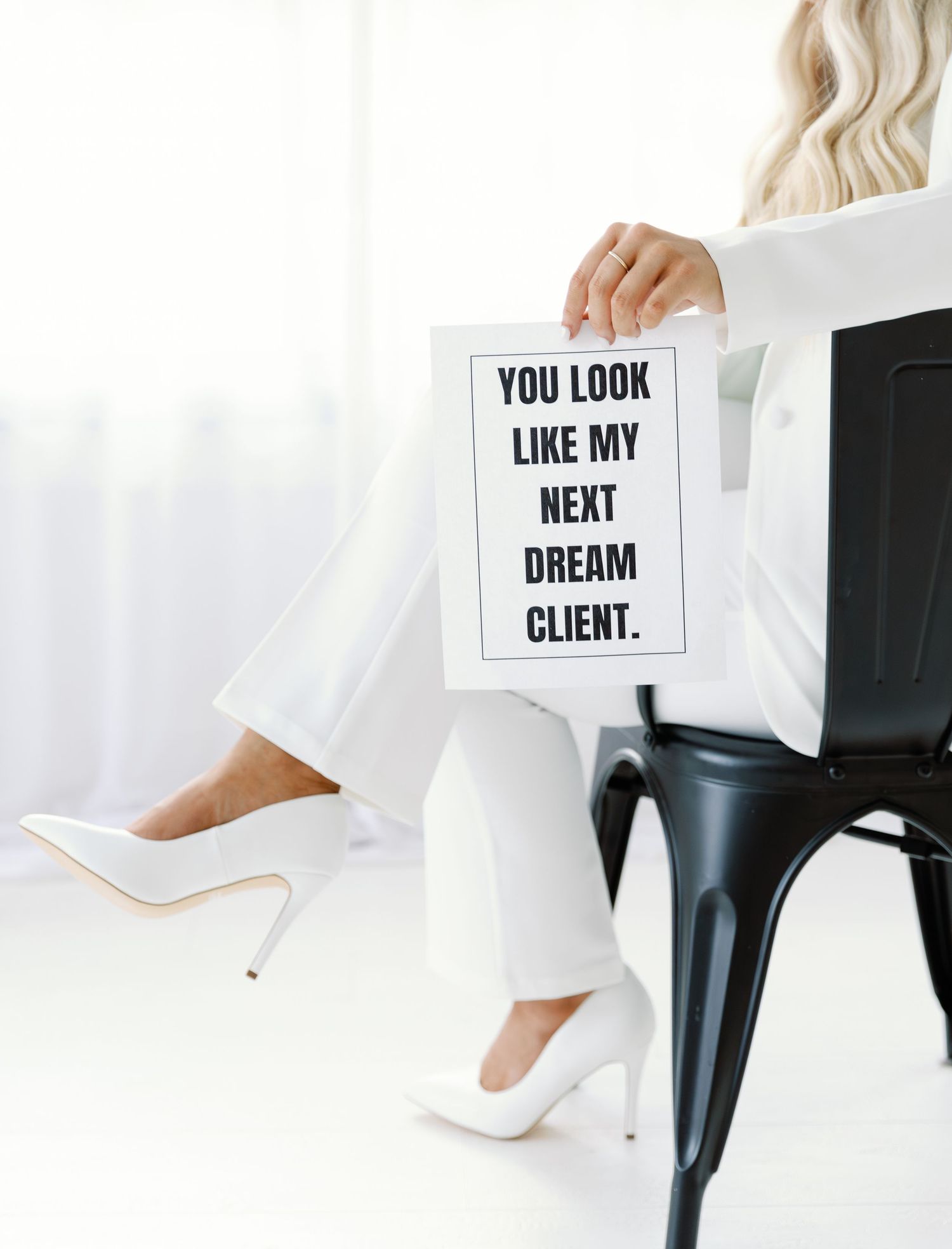 Professional woman in white outfit holding inspirational sign about dream clients while seated with high heels and black chair.