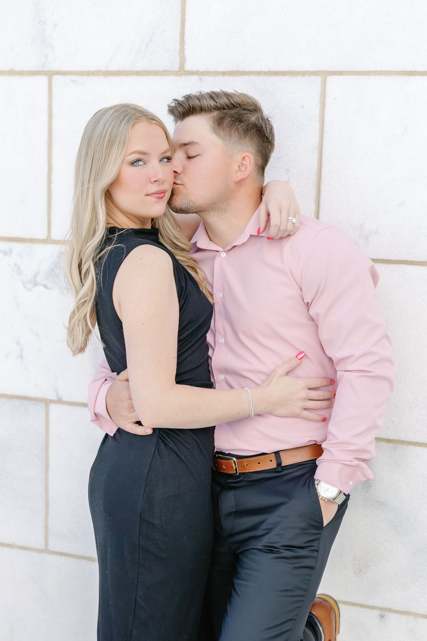 Romantic couple in formal attire embracing against white wall, wearing pink shirt and black dress.