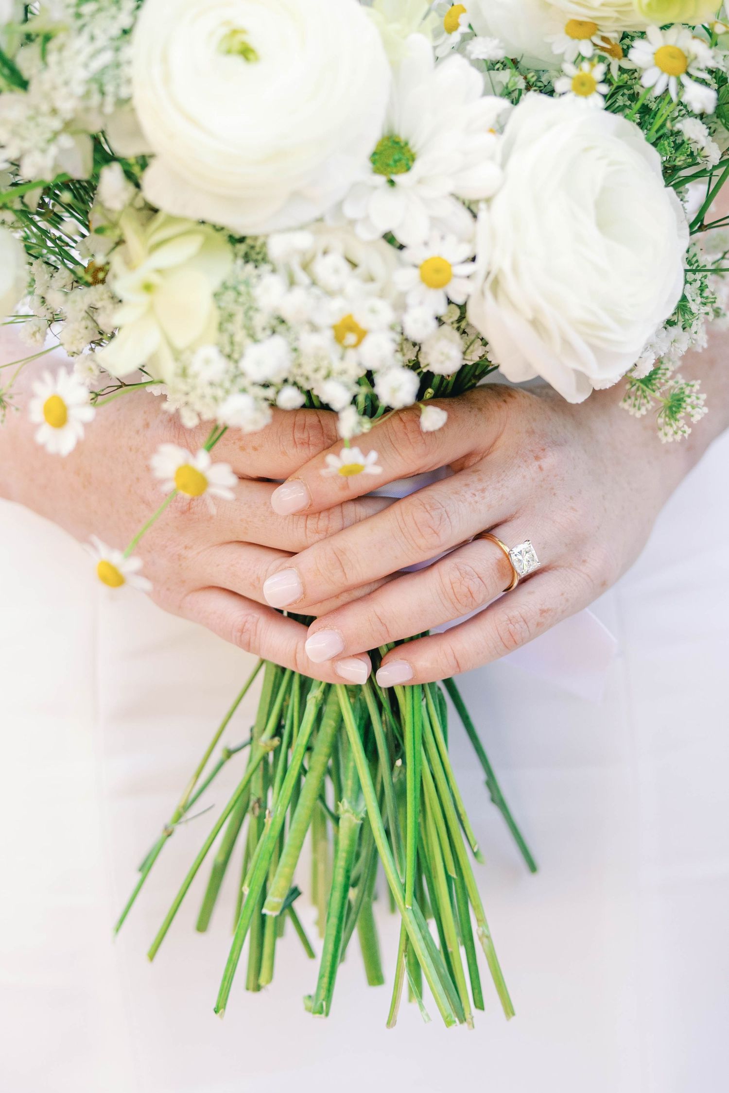 Close-up of hands holding a white and daisy bouquet with a wedding ring displayed.