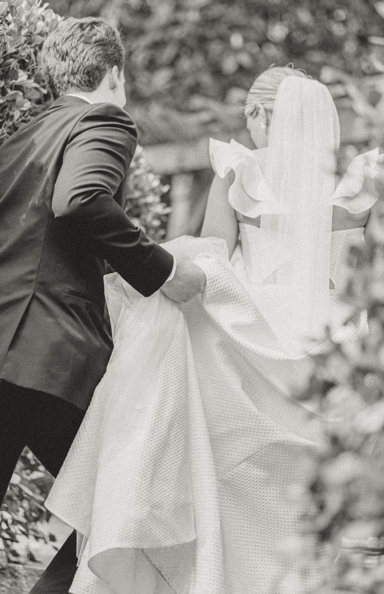 A black and white photograph of a couple outdoors at their wedding, with the groom helping to carry the bride's dress.