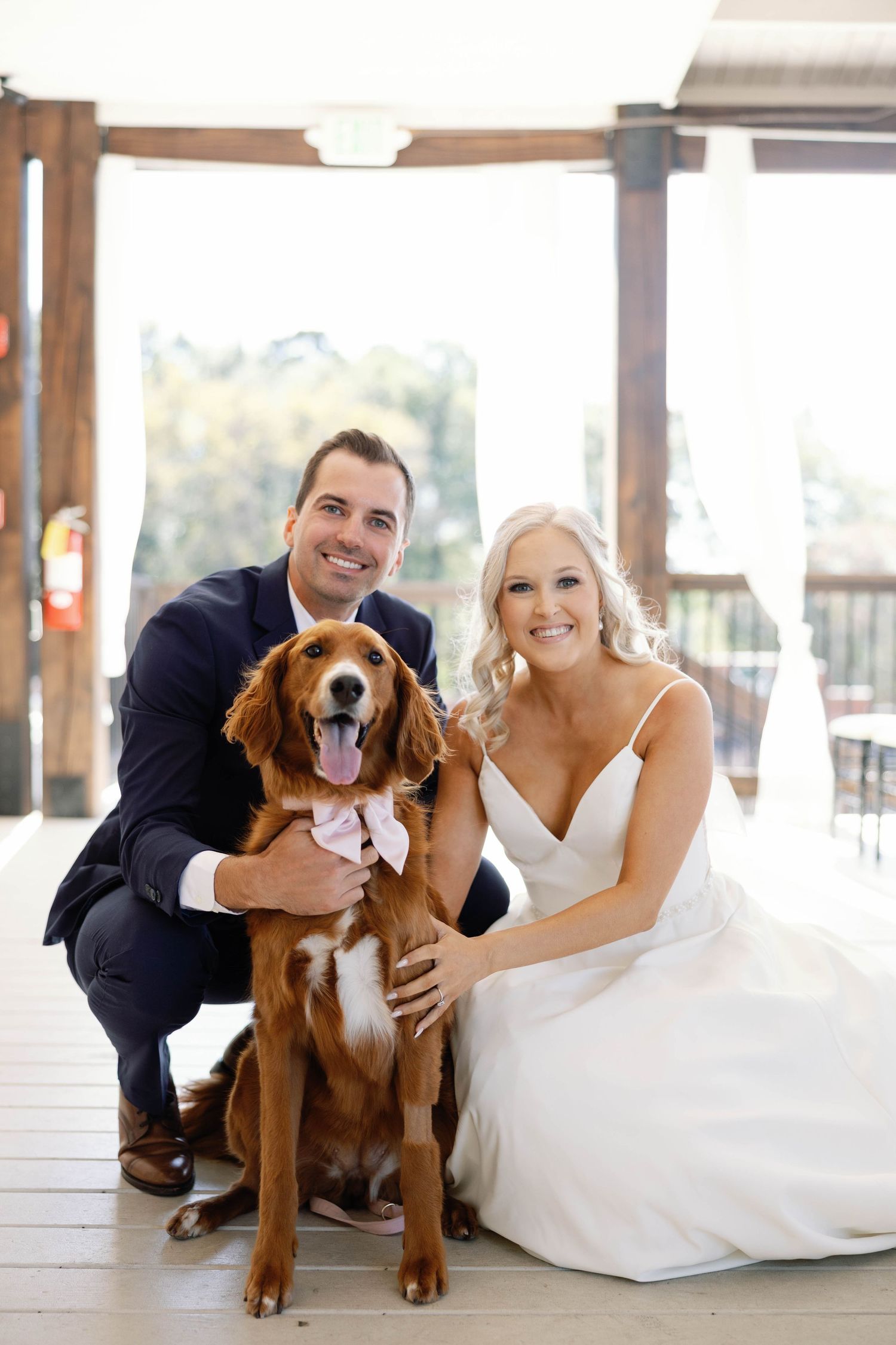 A wedding couple poses with their happy brown and white dog on a covered outdoor porch.