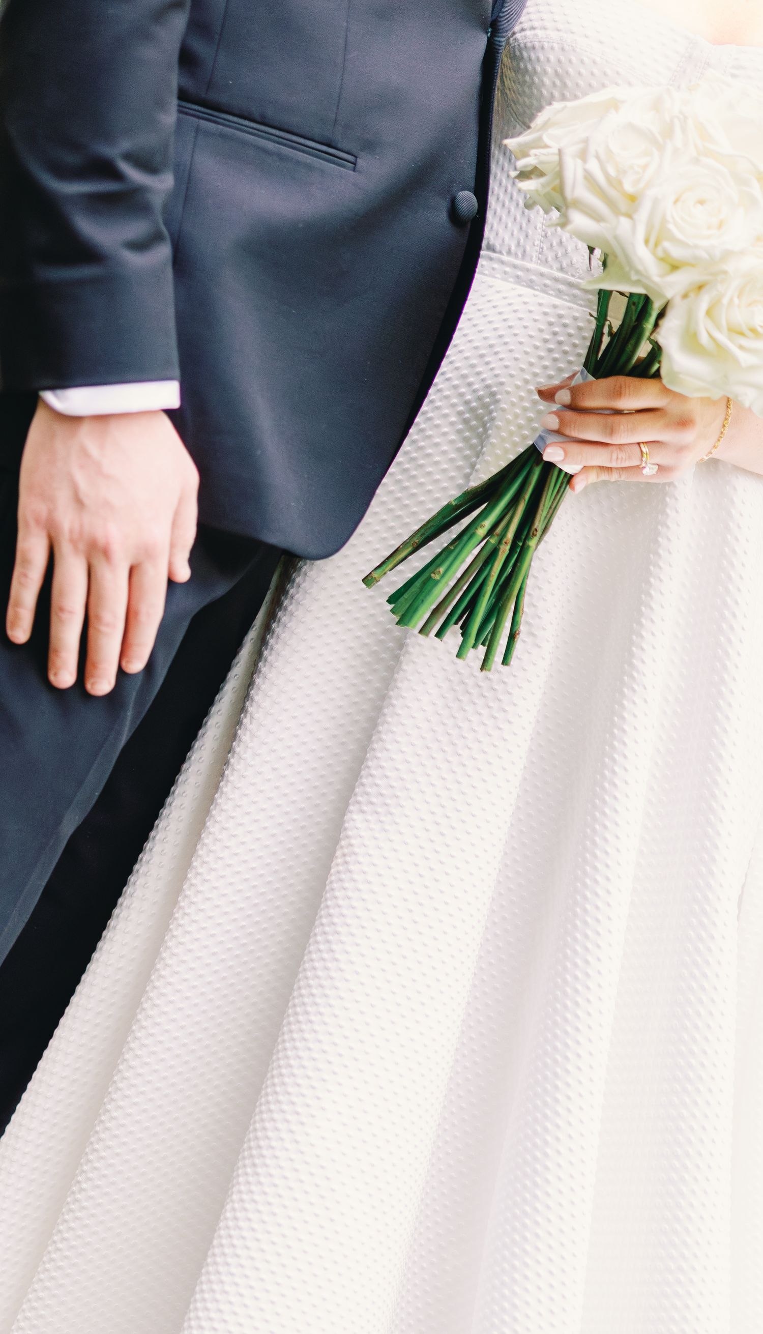 Close-up detail of a navy suit next to a textured white wedding dress with someone holding a bouquet of white roses.