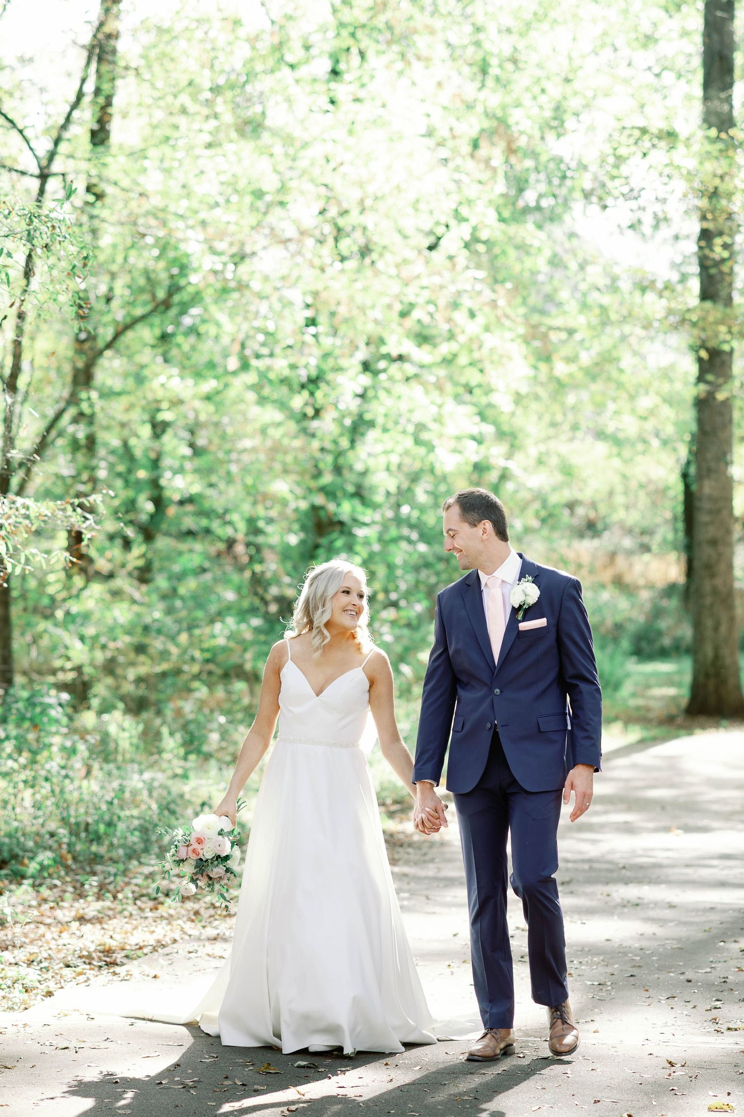 Couple in wedding attire walking and holding hands along a sunlit tree-lined path in a romantic outdoor setting.