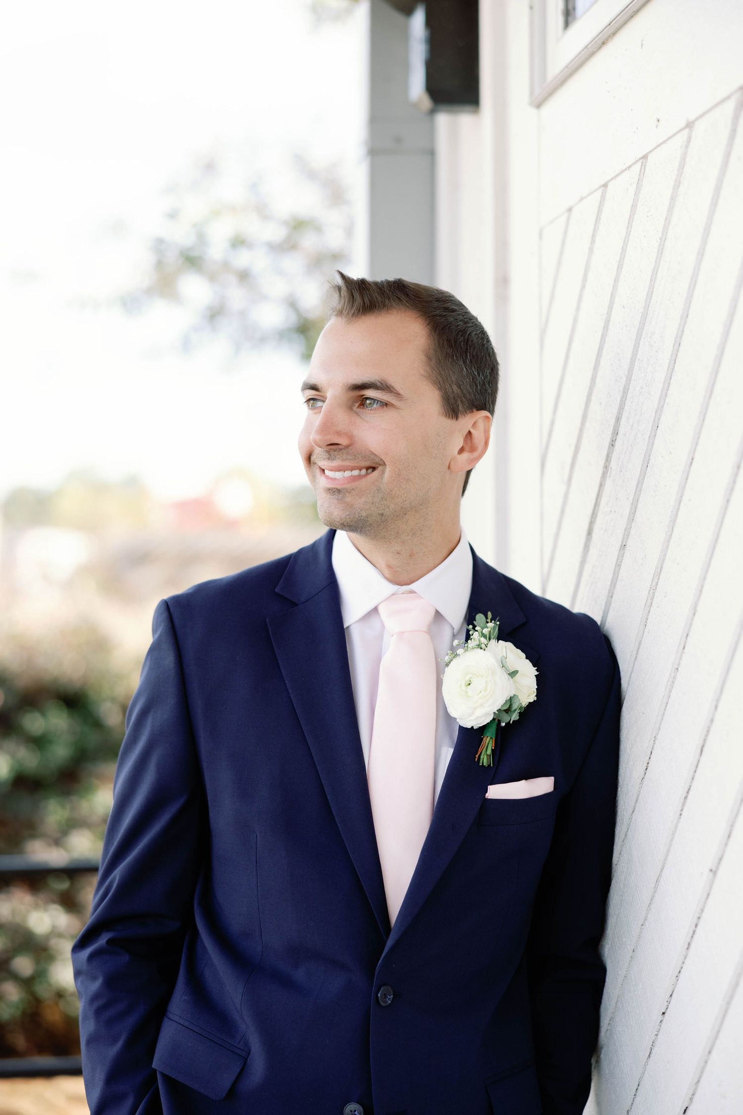 A groom in a navy suit with pink tie and white boutonniere smiles while standing against a white wall.