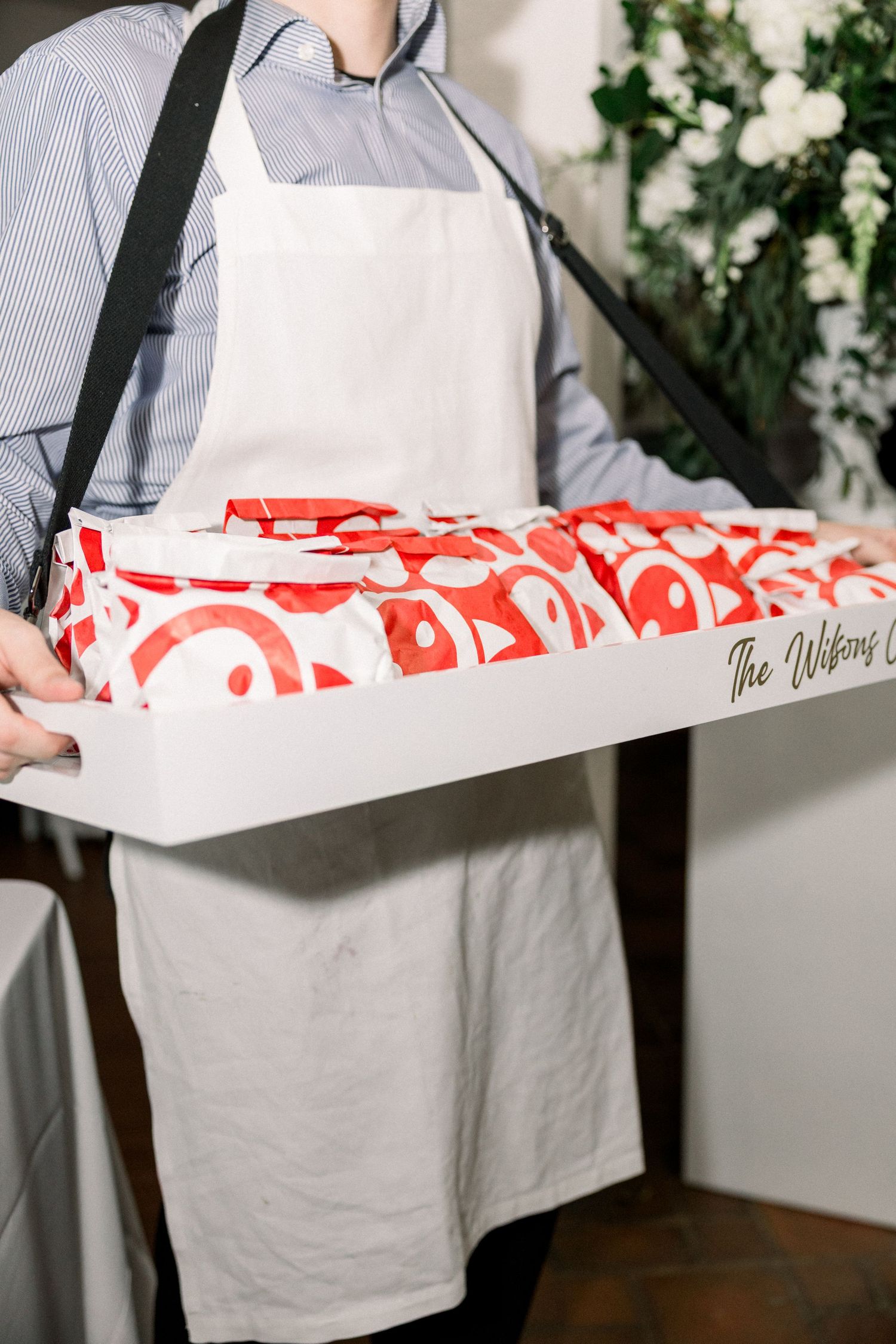 Server wearing white apron carries a tray full of red and white takeout containers at a catering event.