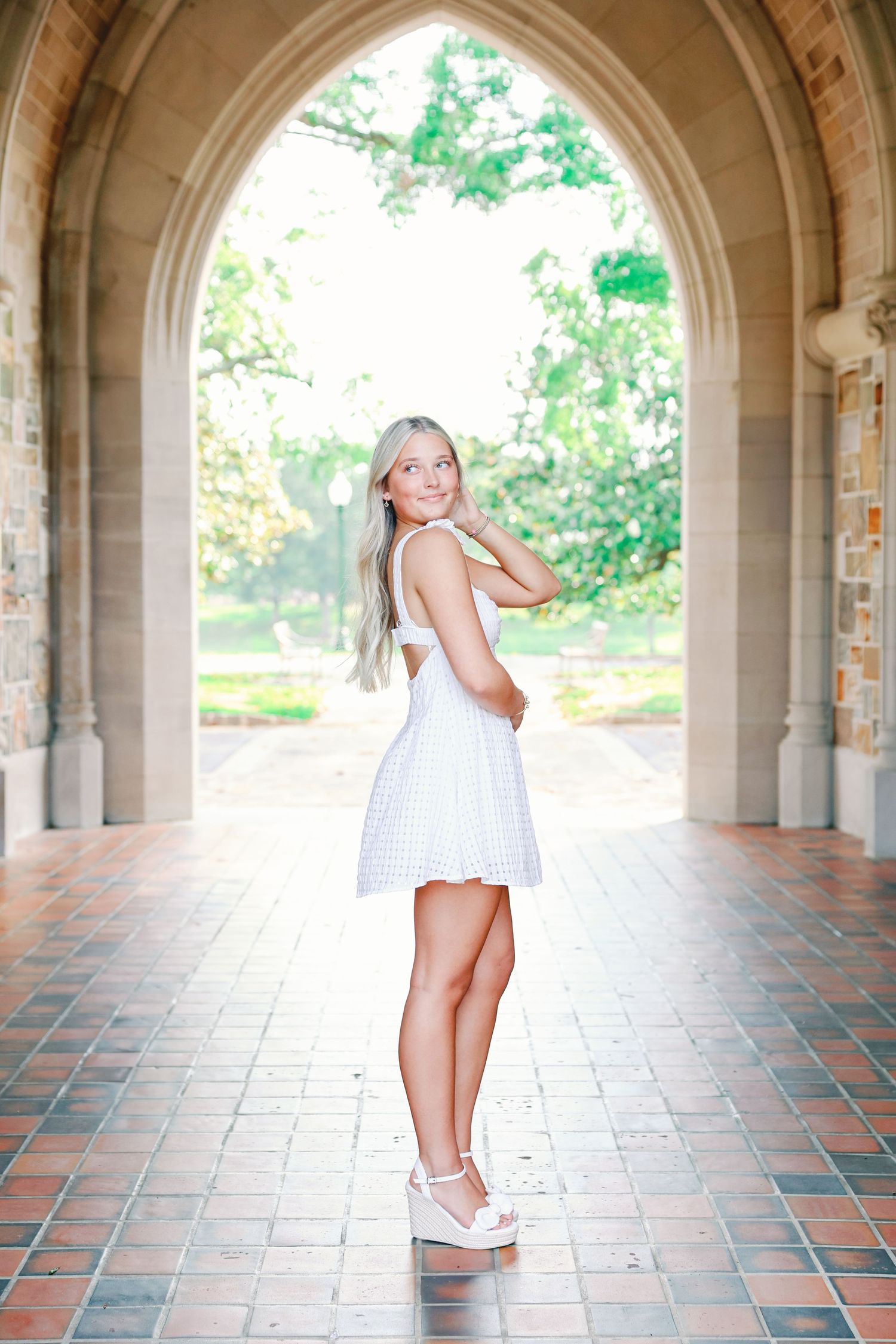 A person in a white sundress poses under a stone archway on a tiled walkway on a bright summer day.