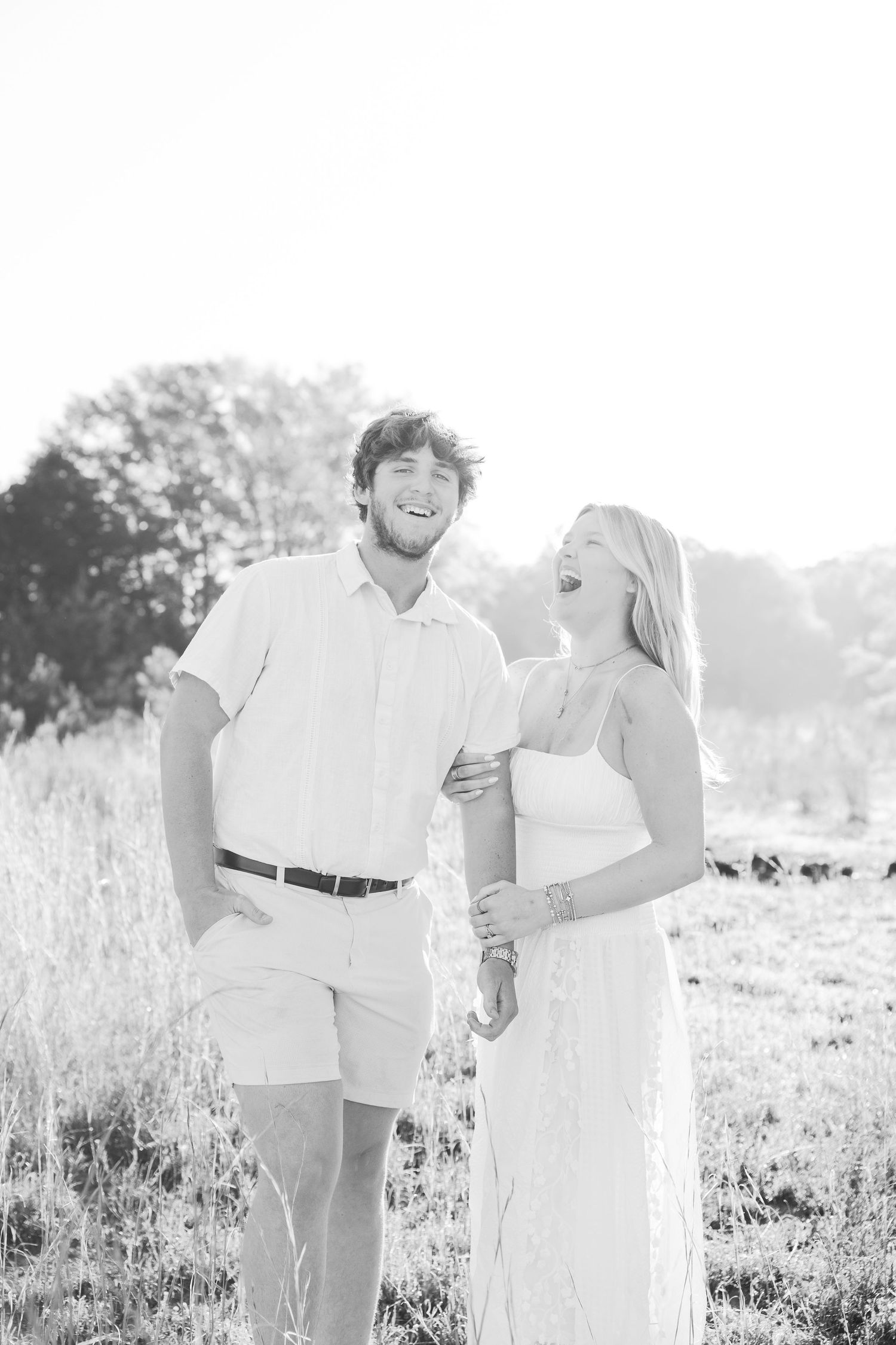 Black and white photo of couple laughing together in sunlit field wearing white outfits.