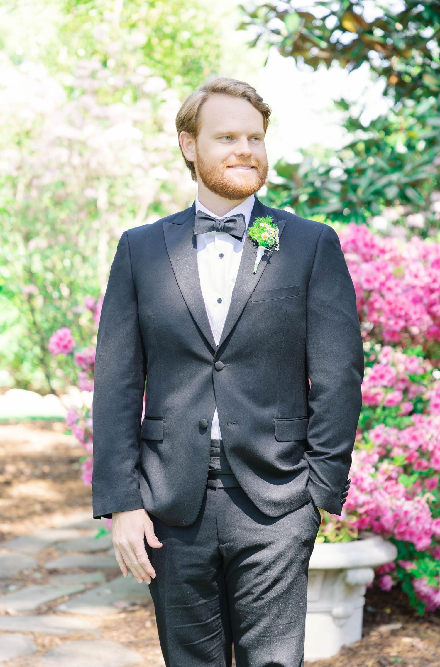 Groom in classic black tuxedo and bowtie poses in garden with pink azaleas in background.