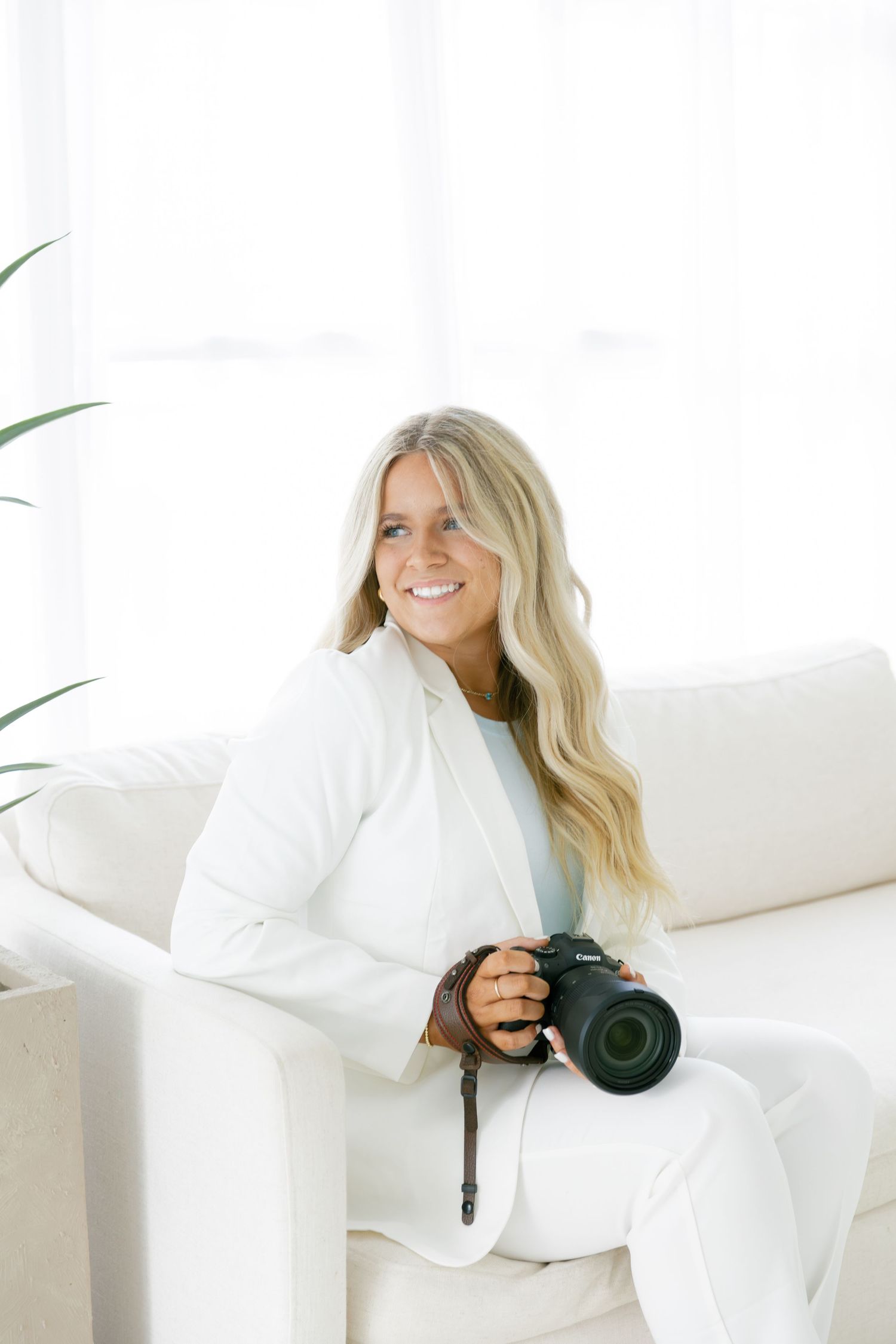 Professional photographer in white outfit sits comfortably on couch while holding camera in bright studio setting.