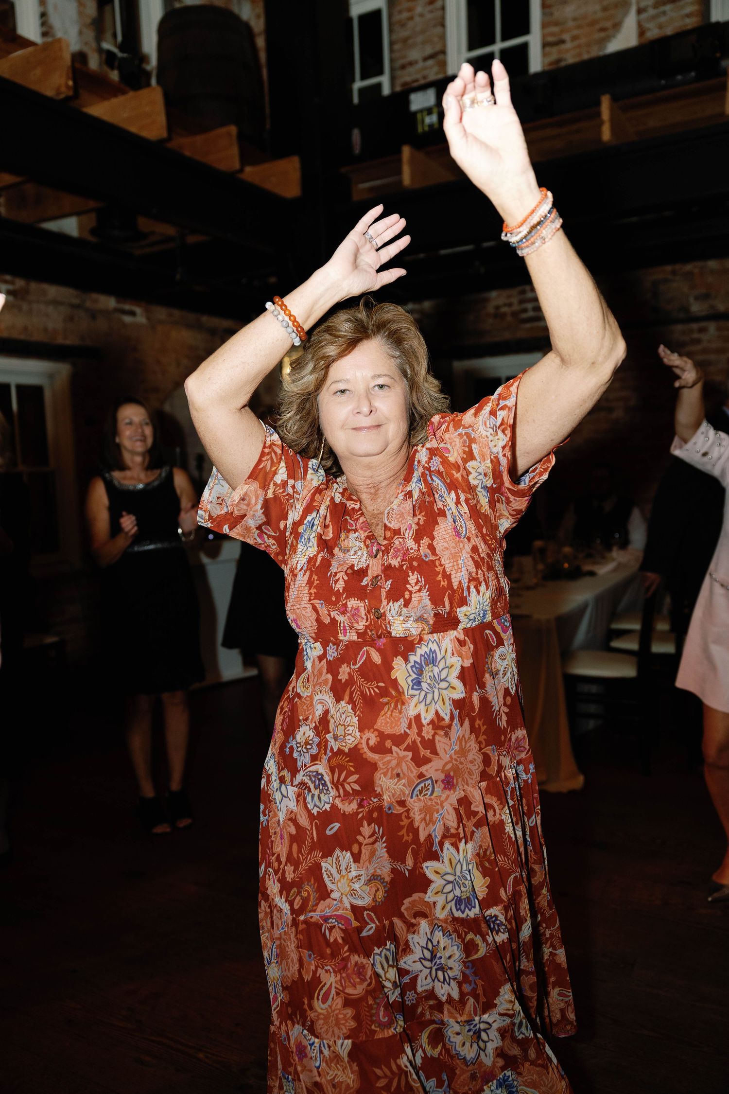 A person in a red floral dress dances with raised arms at an indoor venue with exposed brick walls.