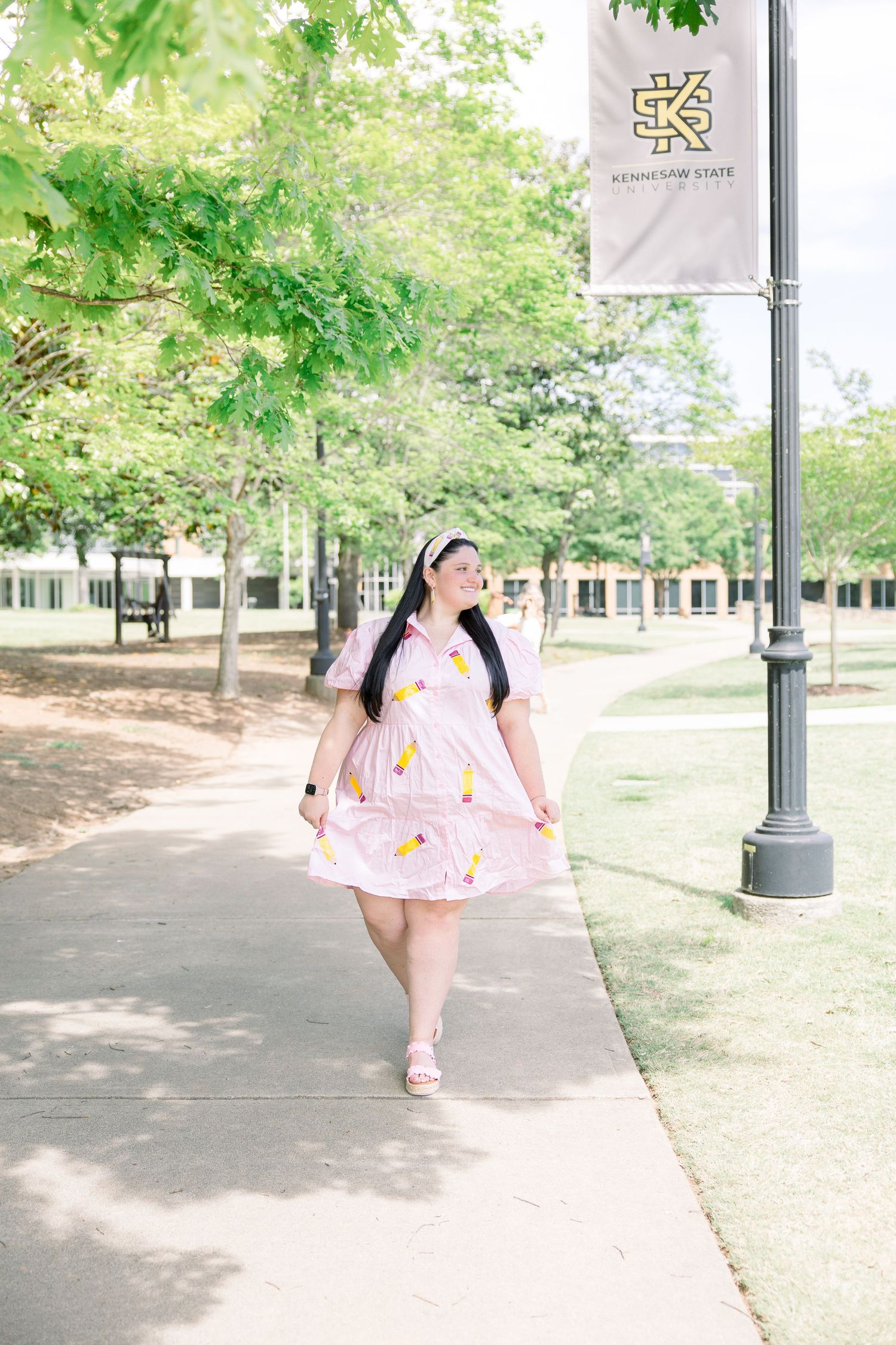 Person in pink pencil-print dress walking on campus sidewalk near university banner.