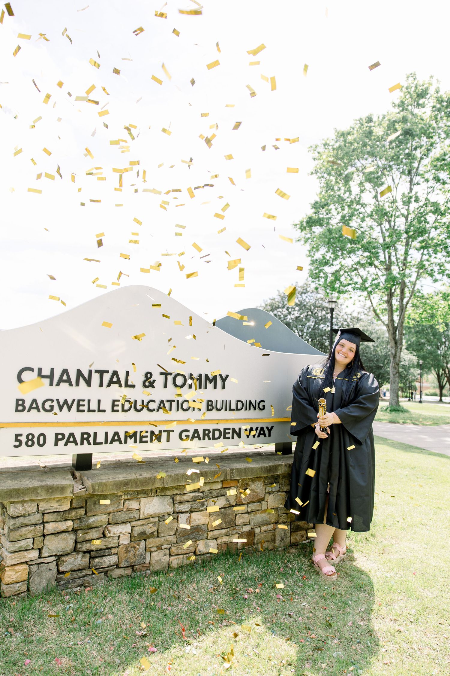 Graduate posing with gold confetti near stone building sign at Bagwell Education Building.