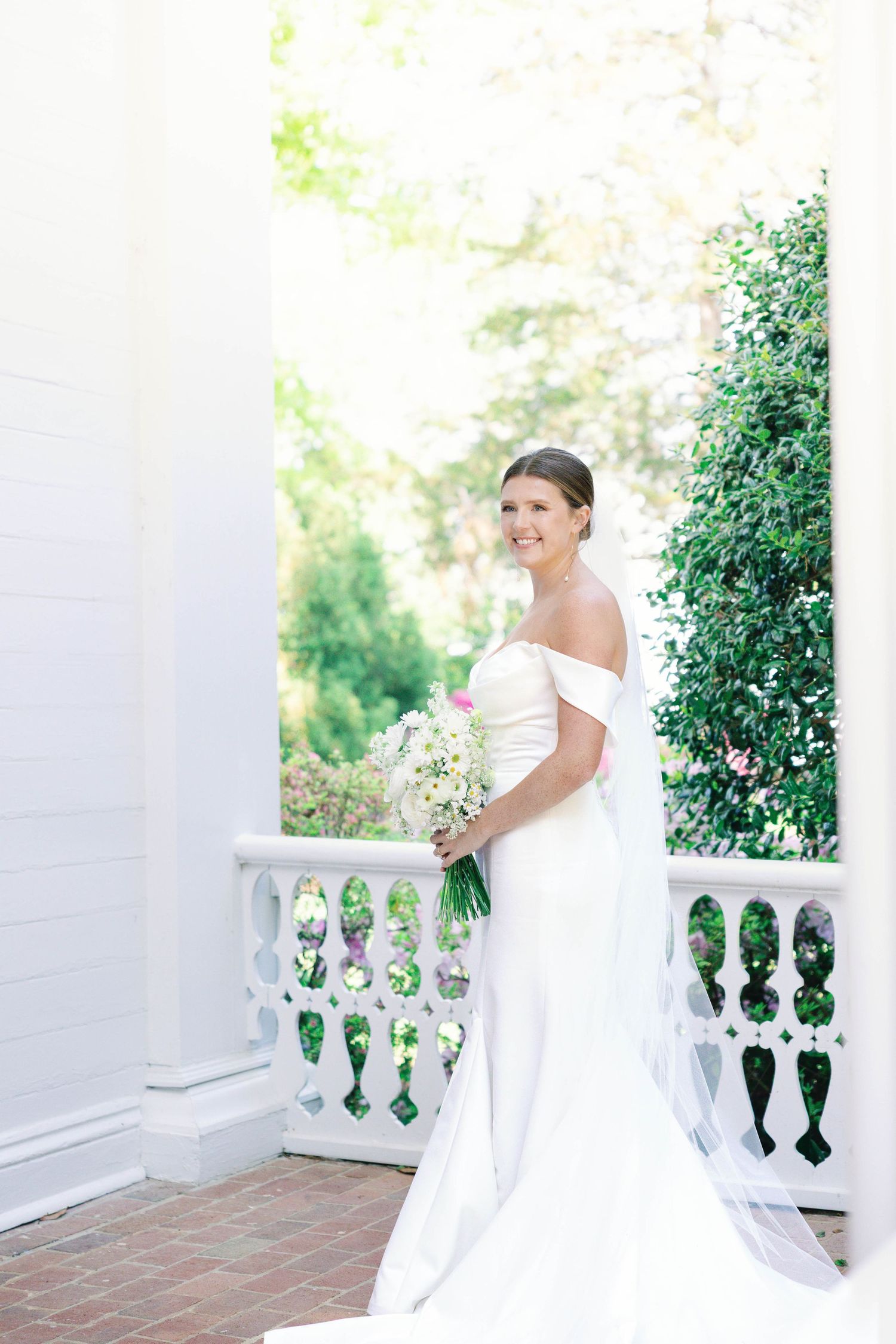 Bride in elegant off-shoulder gown poses on white balustrade porch with bouquet.