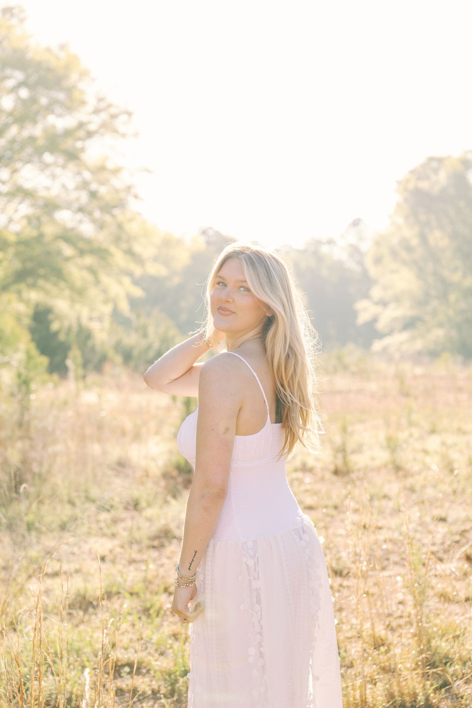Woman in flowing white dress stands in golden sunlit field during sunset.