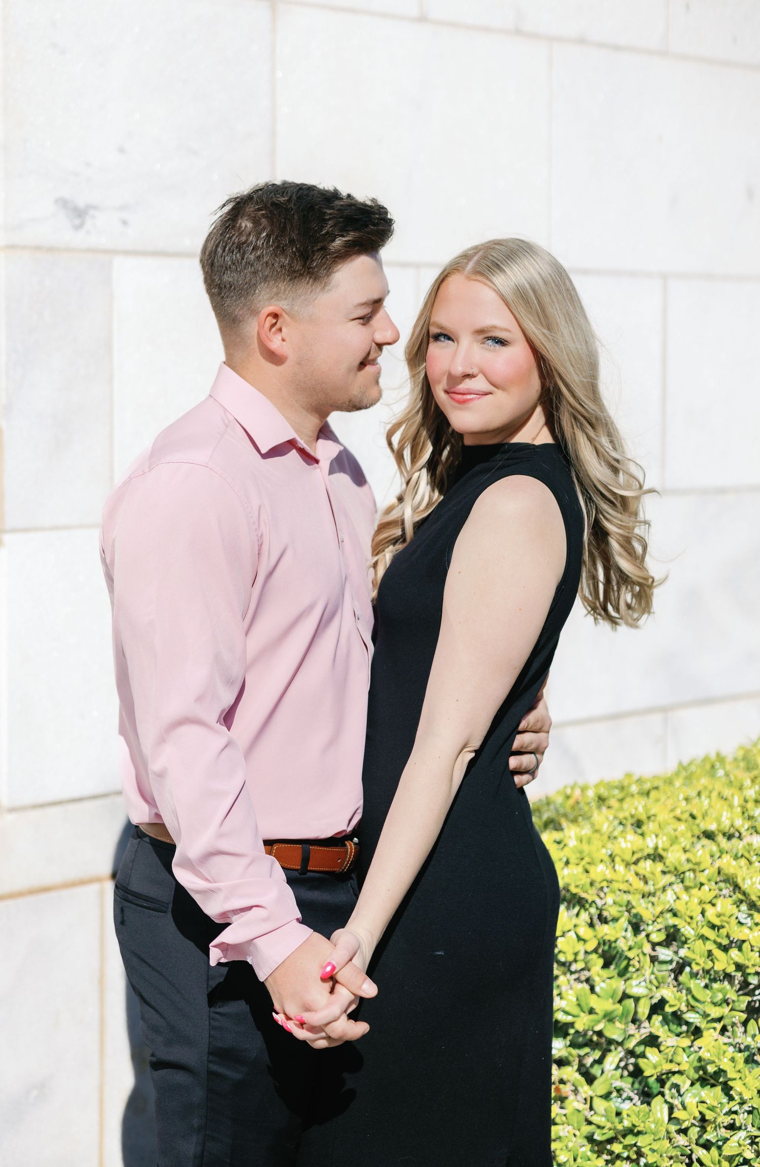 Couple in pink shirt and black dress pose together against white stone wall backdrop.