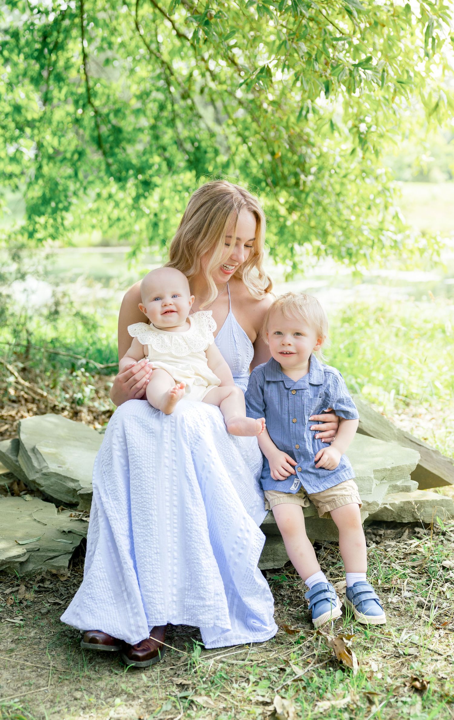 A joyful outdoor family moment captured on rocks under green trees in springtime lighting.