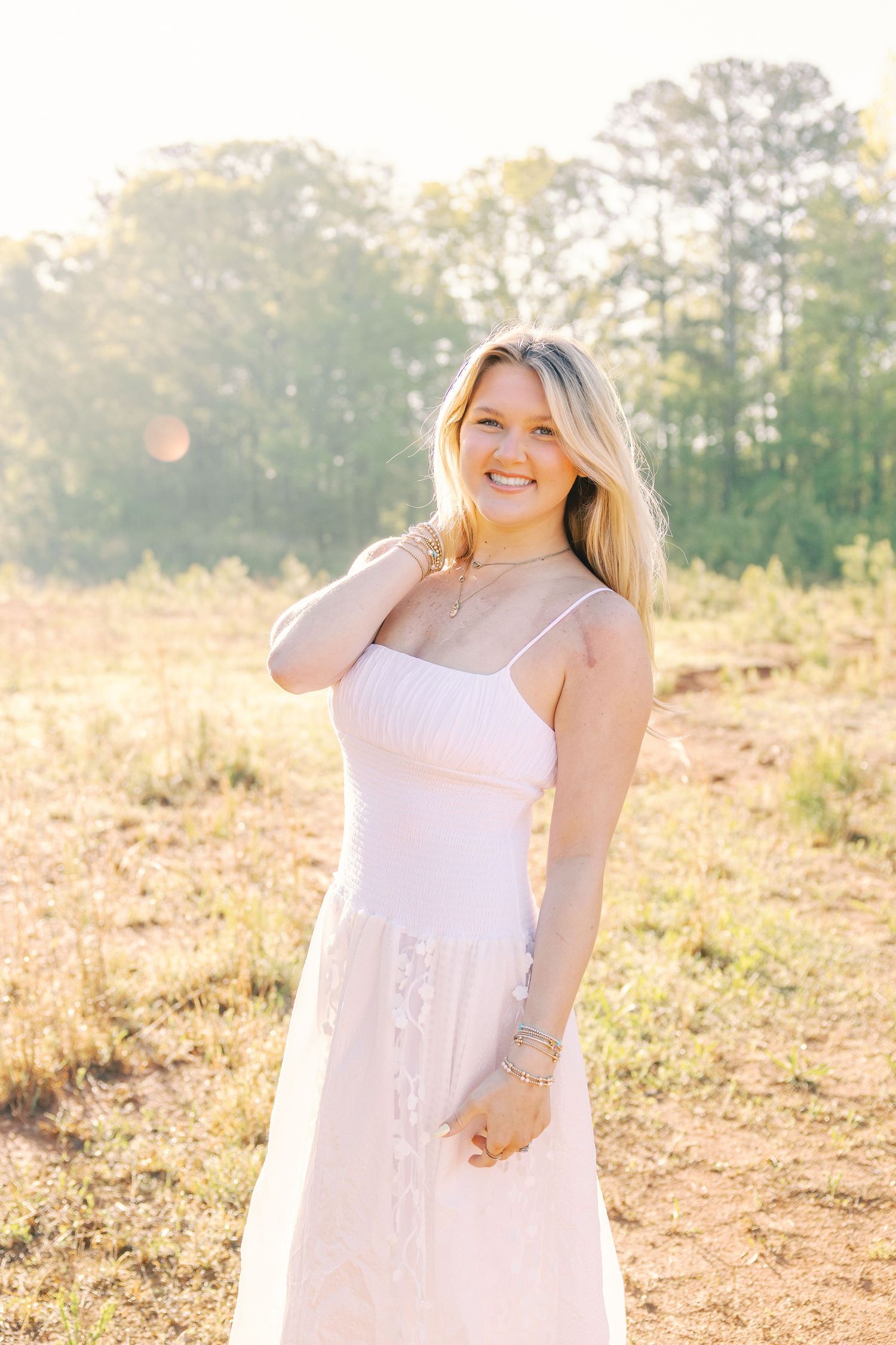 Woman in white dress poses cheerfully in sunlit outdoor setting.