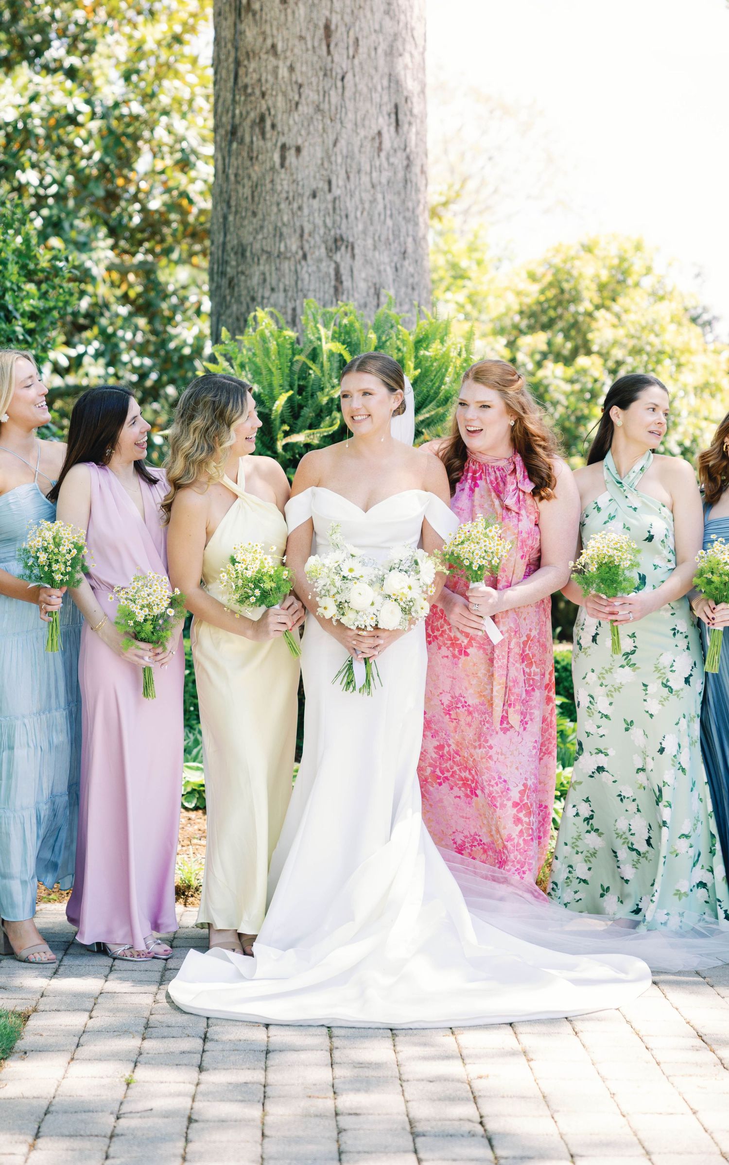 Beautiful bridal party in flowing pastel and floral dresses pose together holding white bouquets on a sunny paved terrace.
