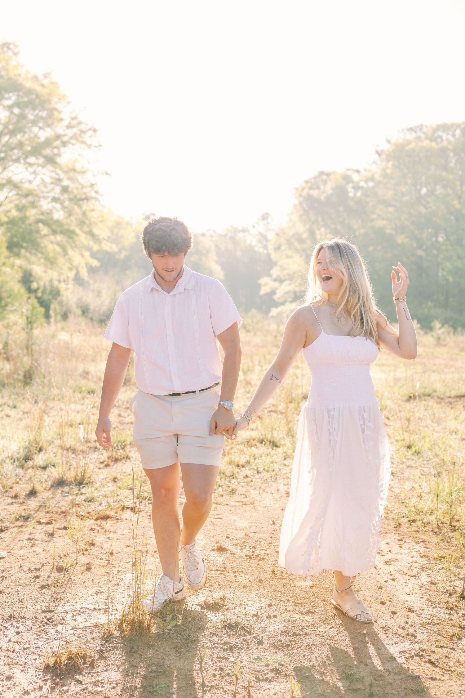 Couple walking hand in hand through sunlit field wearing summer outfits.