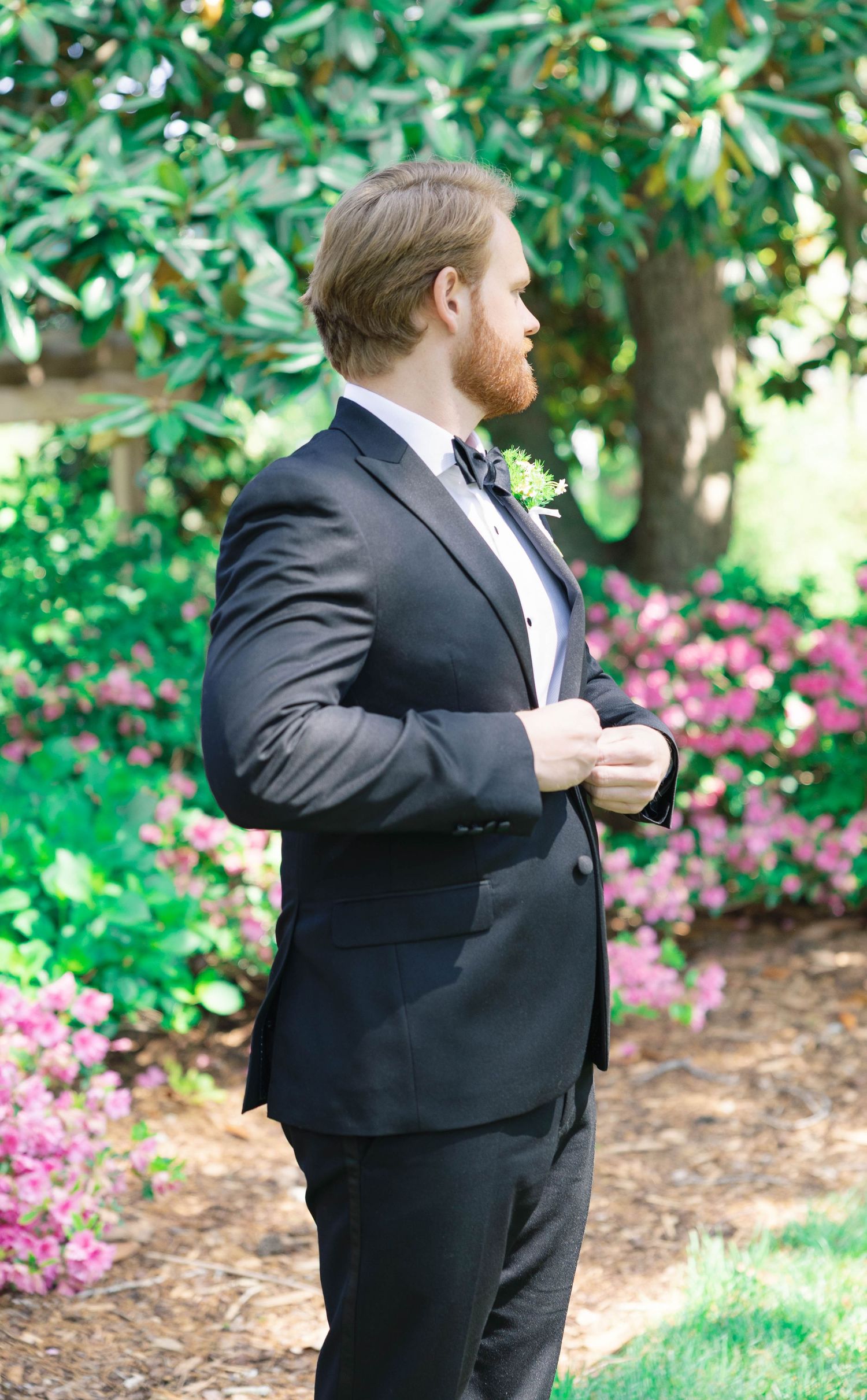 Groom in classic black tuxedo adjusts jacket against garden backdrop.