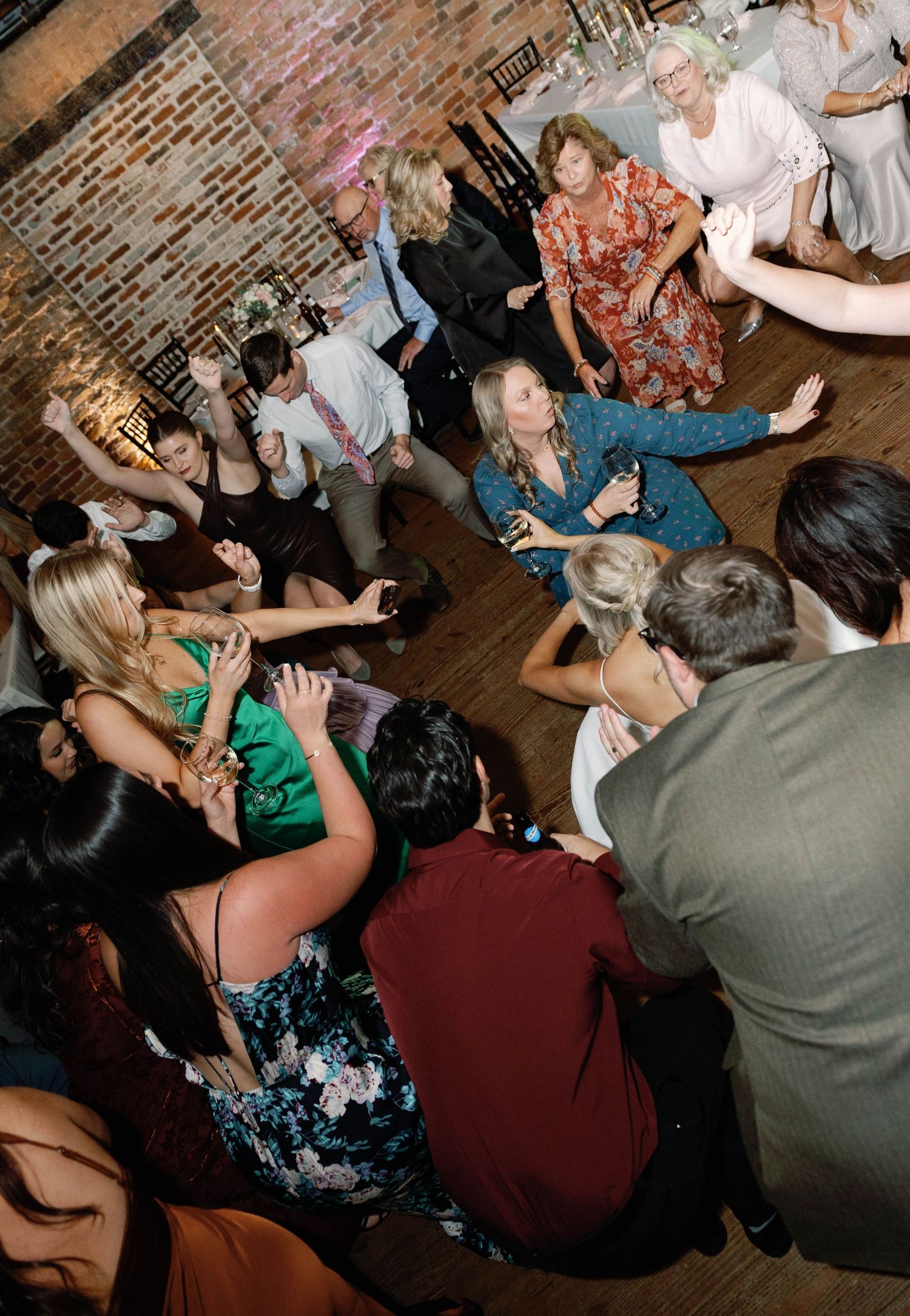 Wedding reception dance floor filled with guests dancing and celebrating in a dimly lit venue with brick walls.