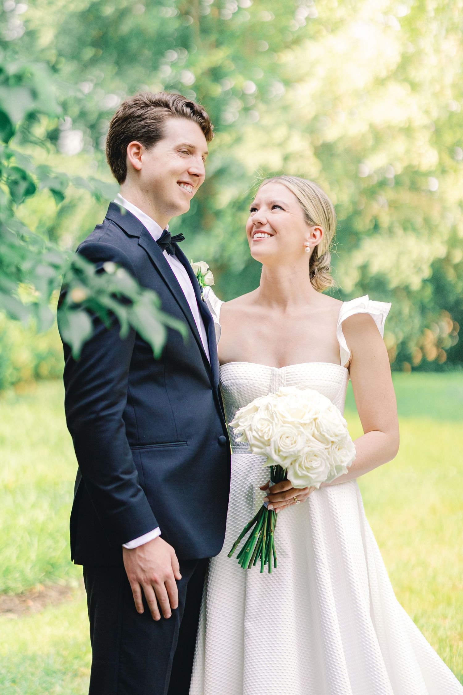 Happy couple sharing a joyful moment on their wedding day in a sun-dappled garden setting with white rose bouquet.
