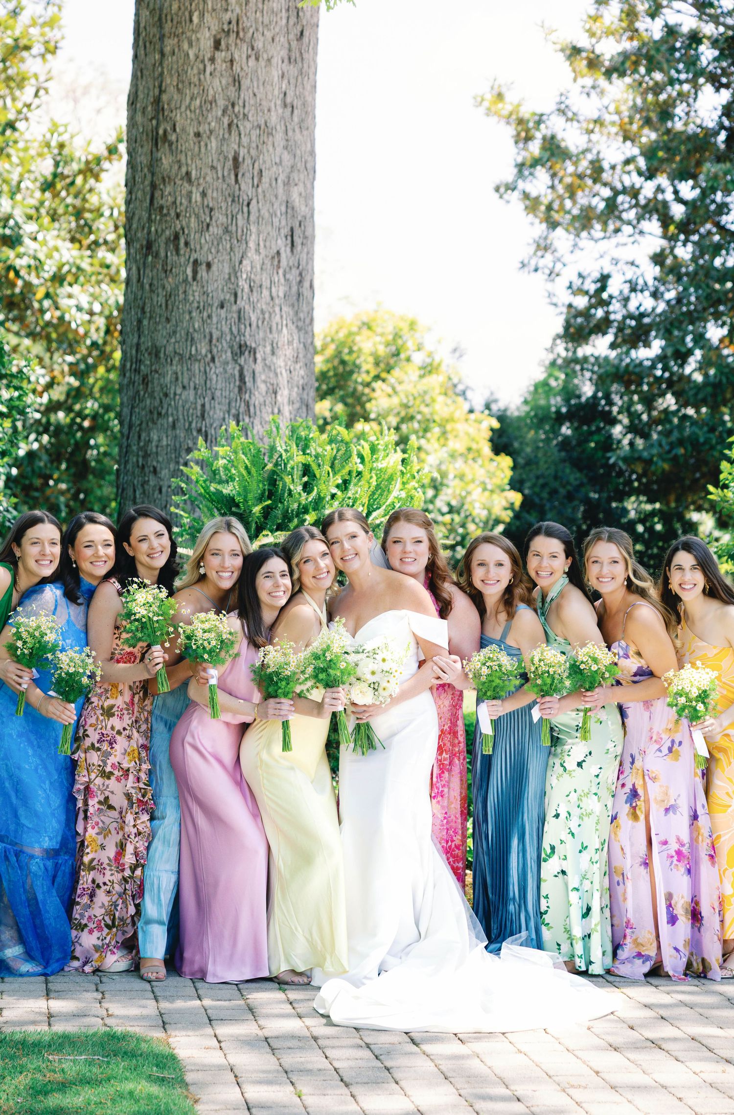 Bridal party in colorful mismatched dresses poses together under large tree.
