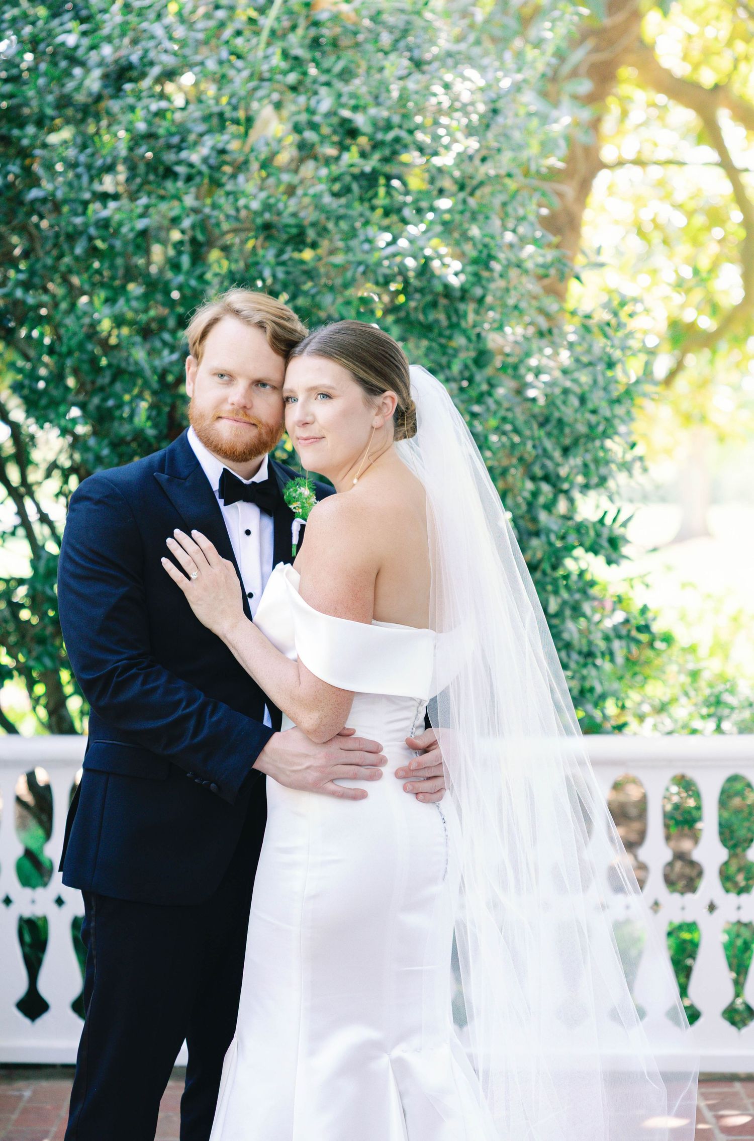 Newlyweds in formal attire embrace on white balcony with garden backdrop.