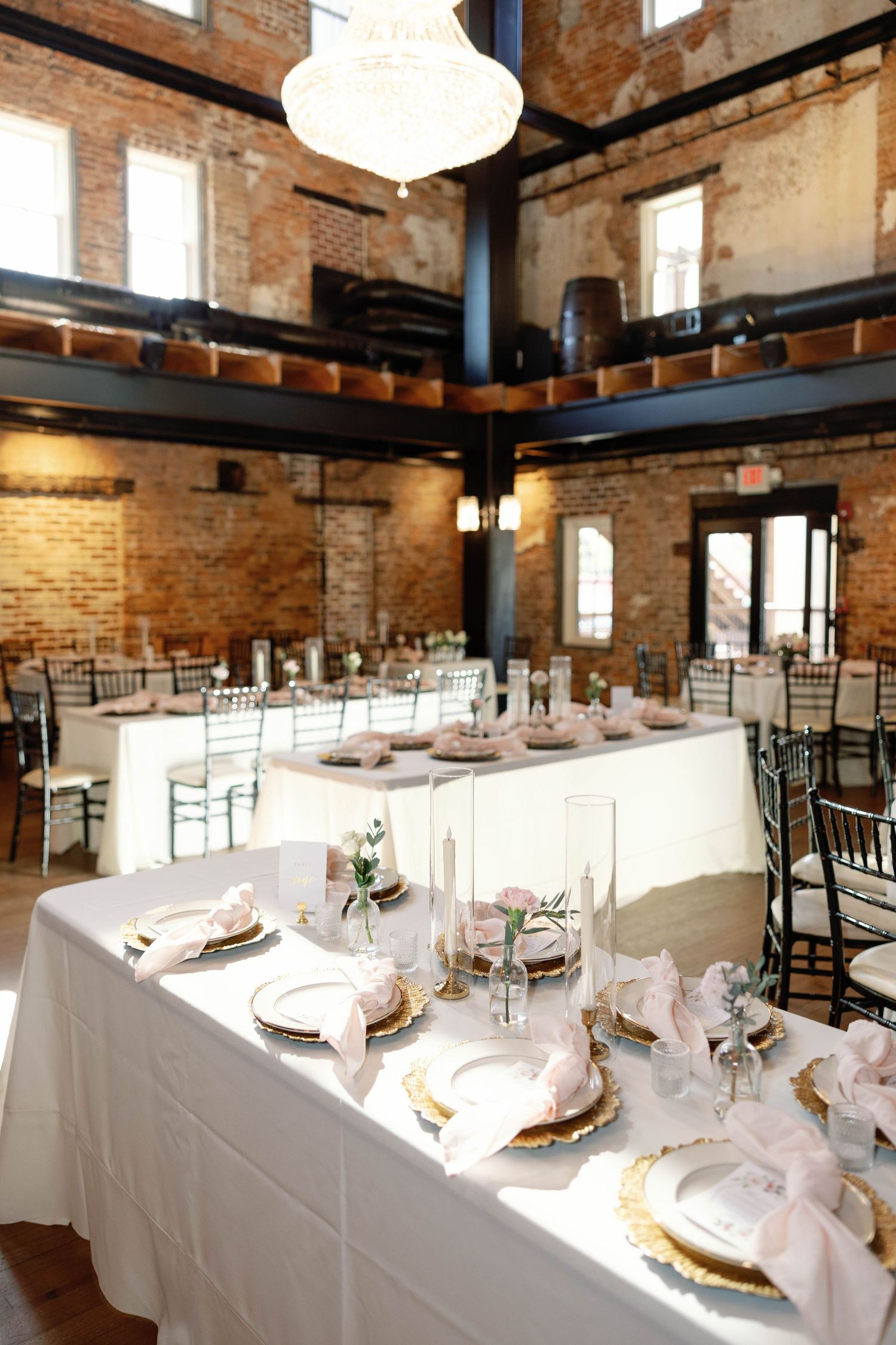 Elegant wedding reception setup in rustic brick venue with white tablecloths and crystal chandeliers.