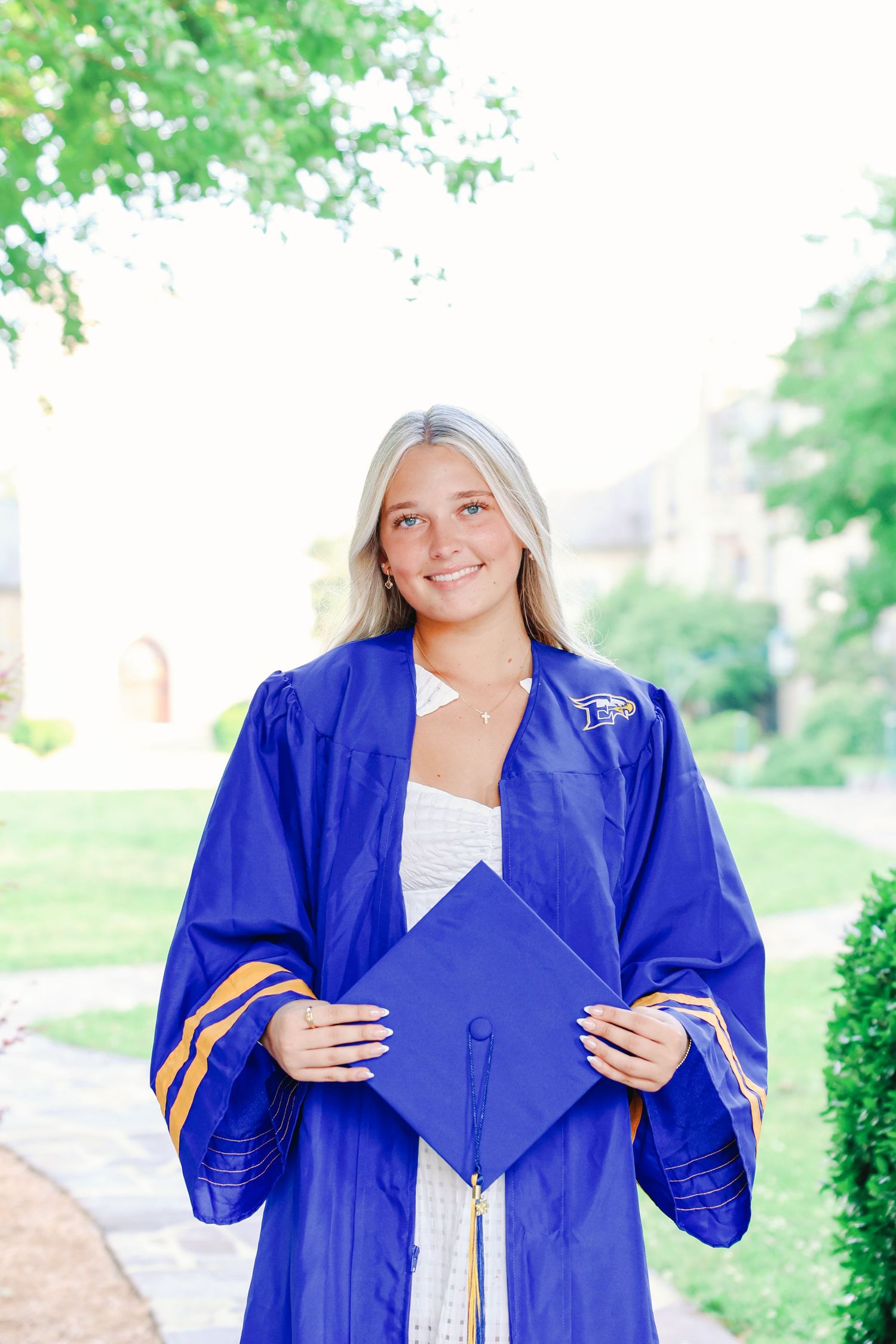 Graduate in royal blue cap and gown holds graduation cap while standing on outdoor path surrounded by greenery.
