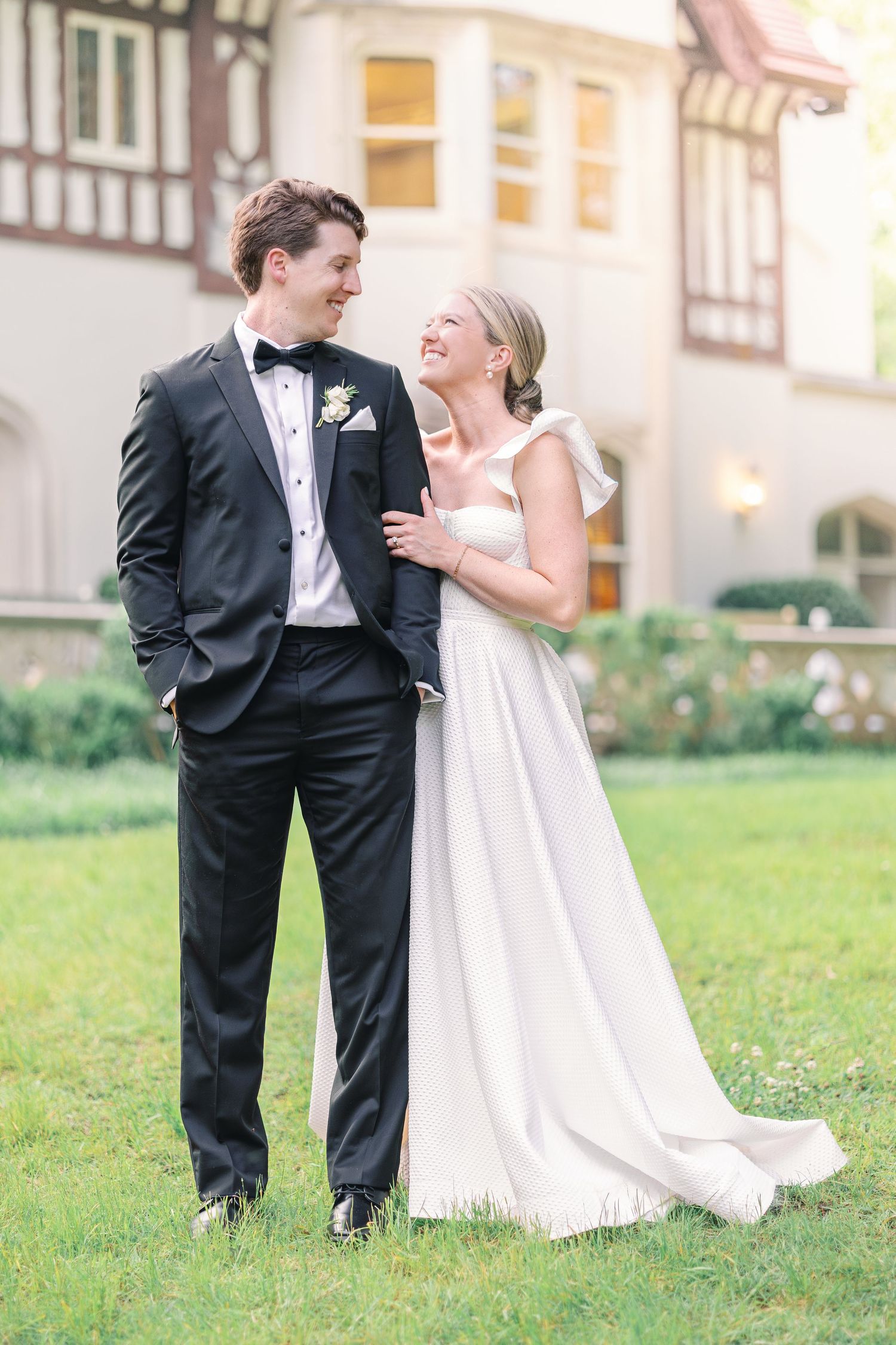 Newlywed couple shares a joyful laugh together on a manicured lawn in front of a stately building.