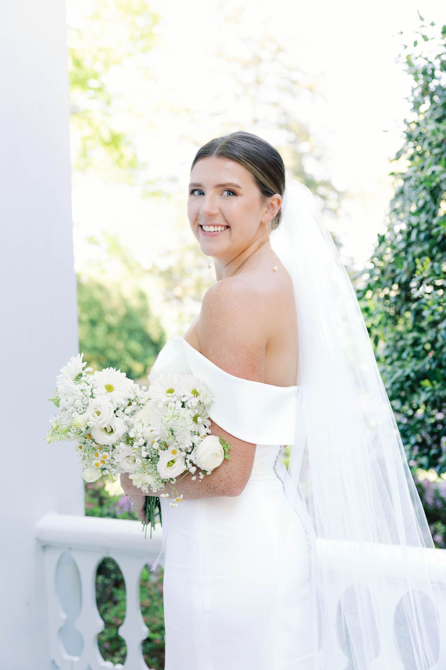 Radiant bride in off-shoulder gown poses with bouquet on white balcony.