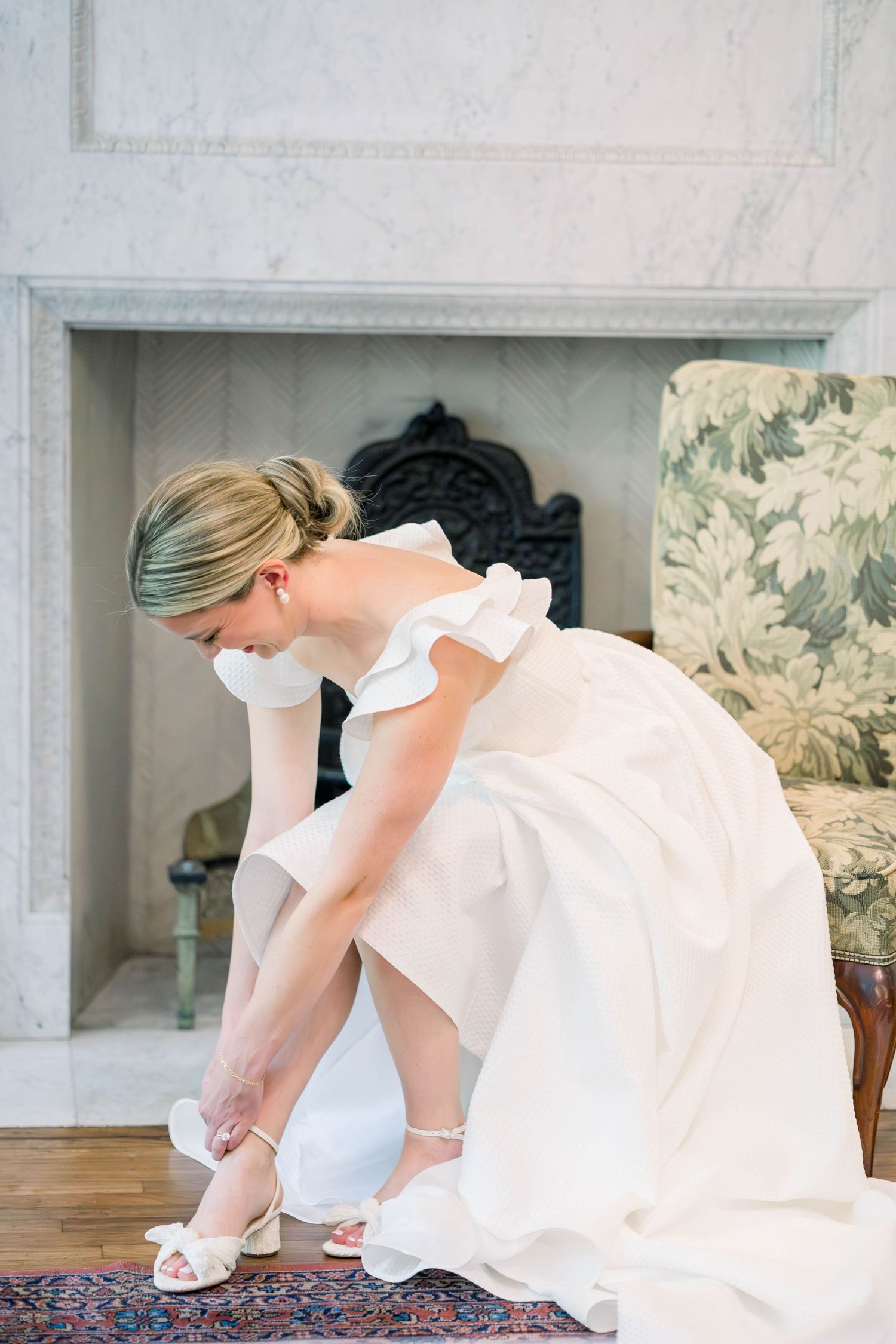 Elegant bride in white gown adjusts her ankle strap sandal while getting ready in a room with ornate fireplace and floral chair.