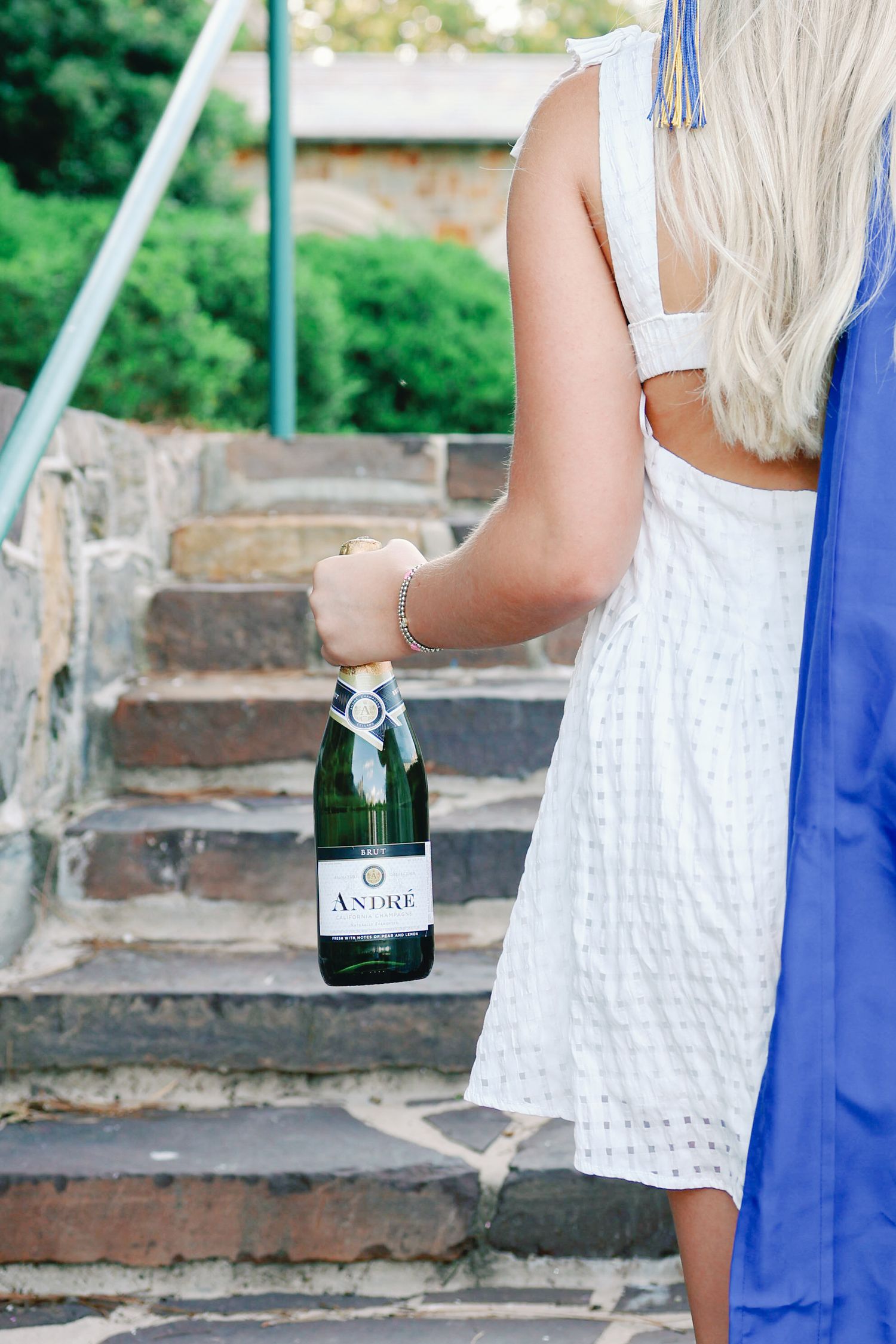 Person in a white dress holding a bottle of wine while standing on stone steps outdoors.