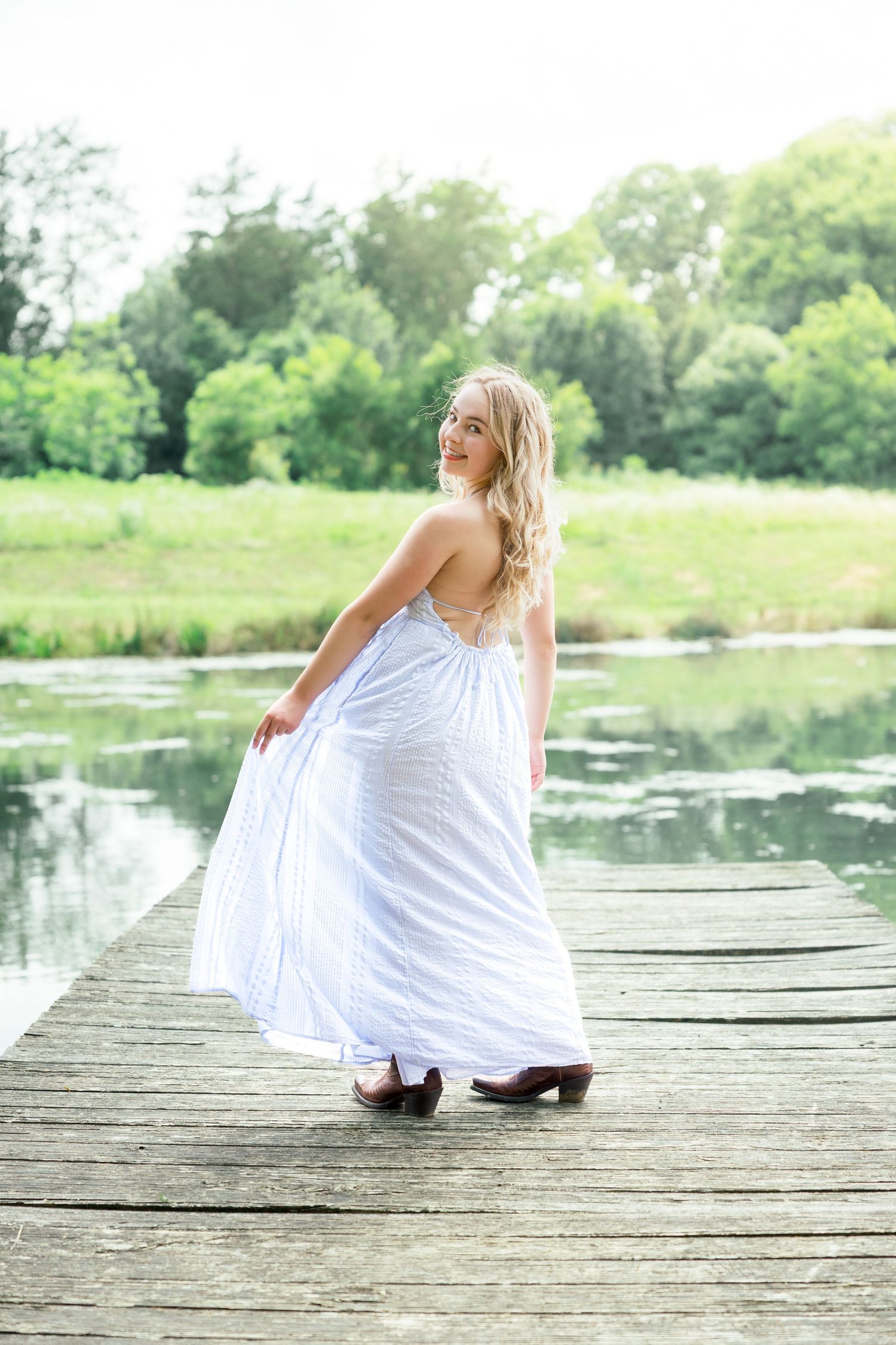 A figure in a flowing white dress stands on a wooden dock overlooking a serene pond surrounded by lush greenery.