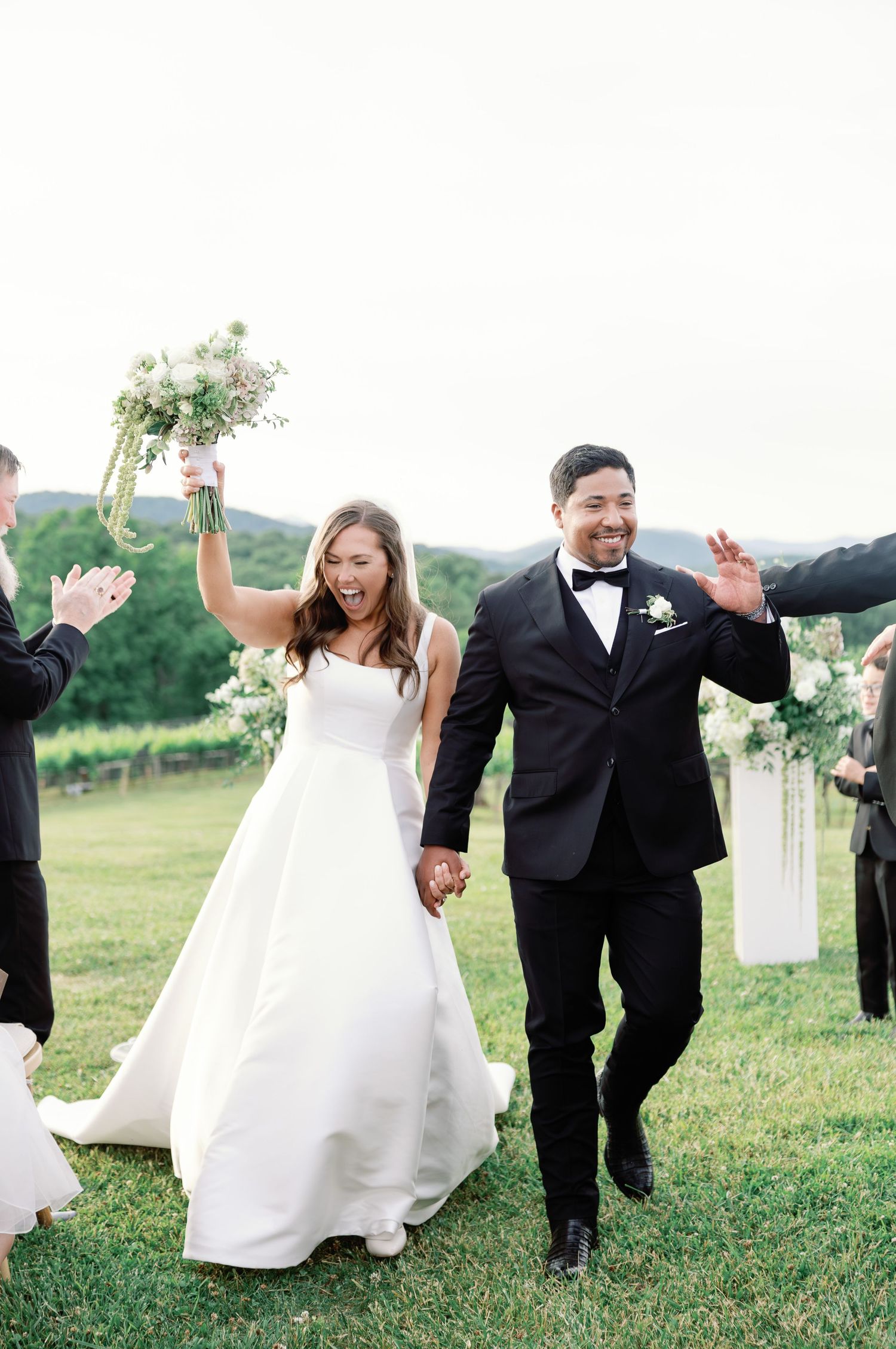 Joyful newlyweds walk down the aisle, bride in white gown holding bouquet aloft, groom in black tuxedo, outdoor wedding.